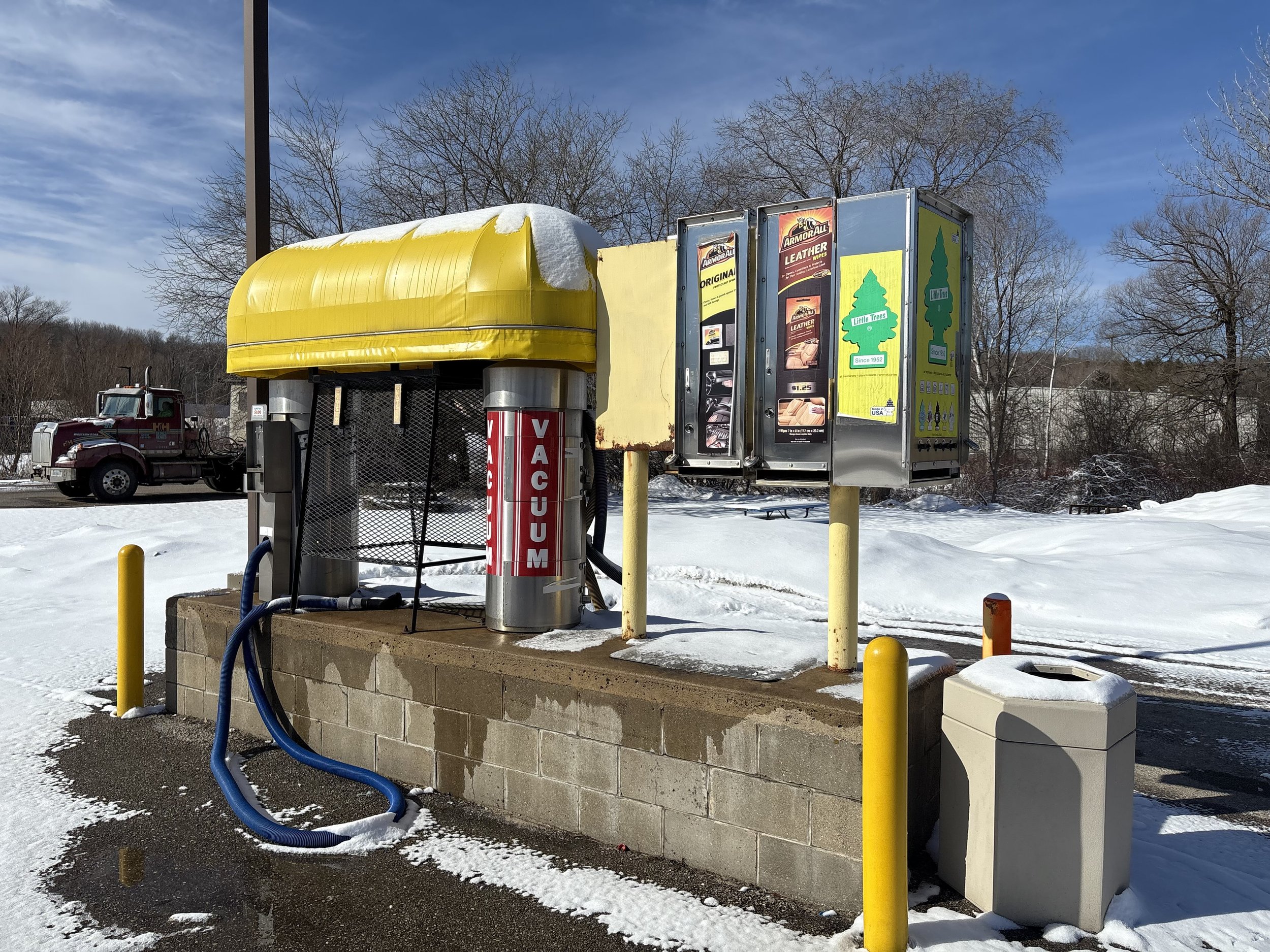 Outdoor car vacuum station with yellow canopy and vending machines for car products, surrounded by snow, with a truck in the background and trees nearby.