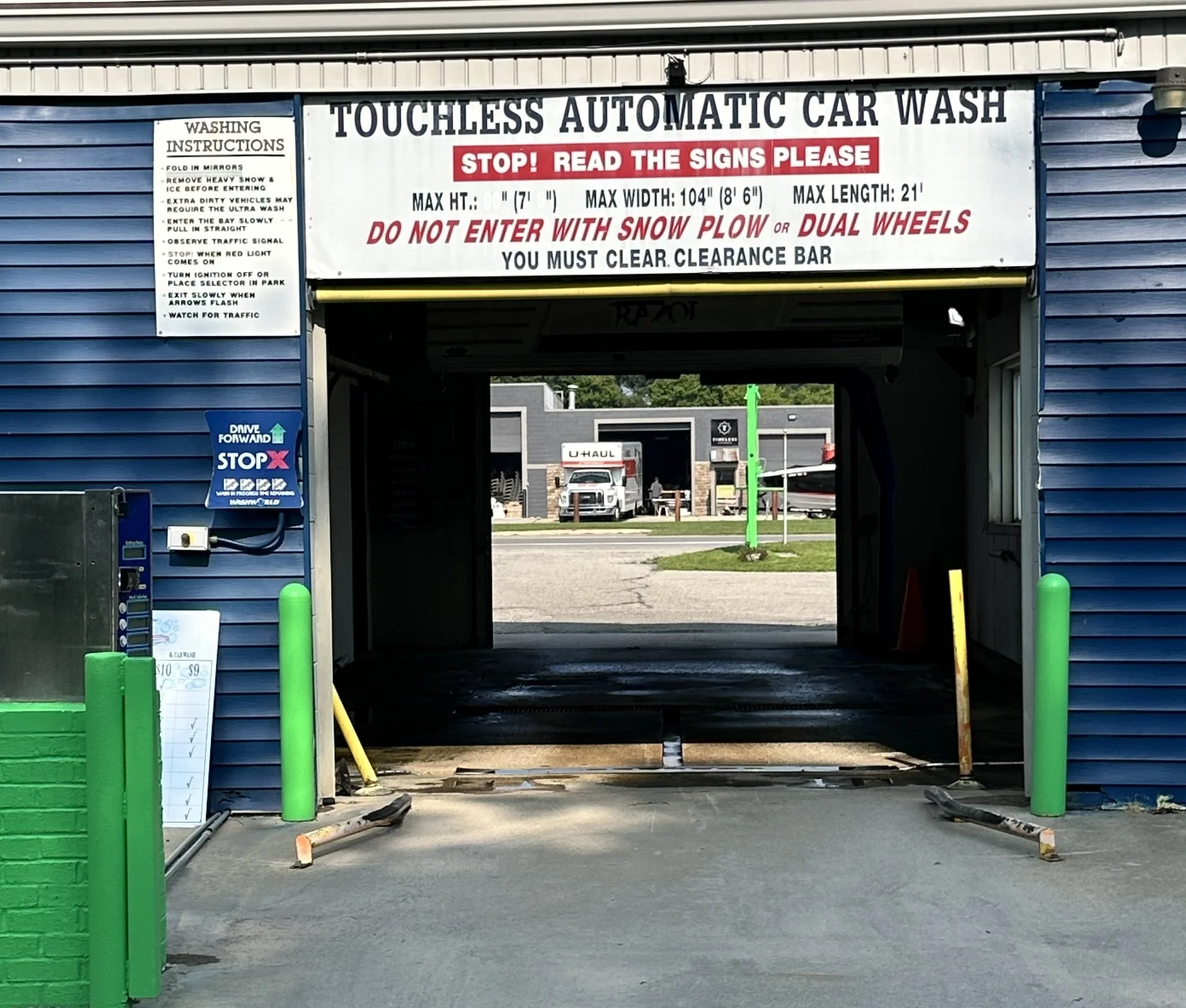 Touchless automatic car wash entrance with signage, including washing instructions, restrictions, and an exterior payment machine. Yellow building with a closed garage door and snowy ground.