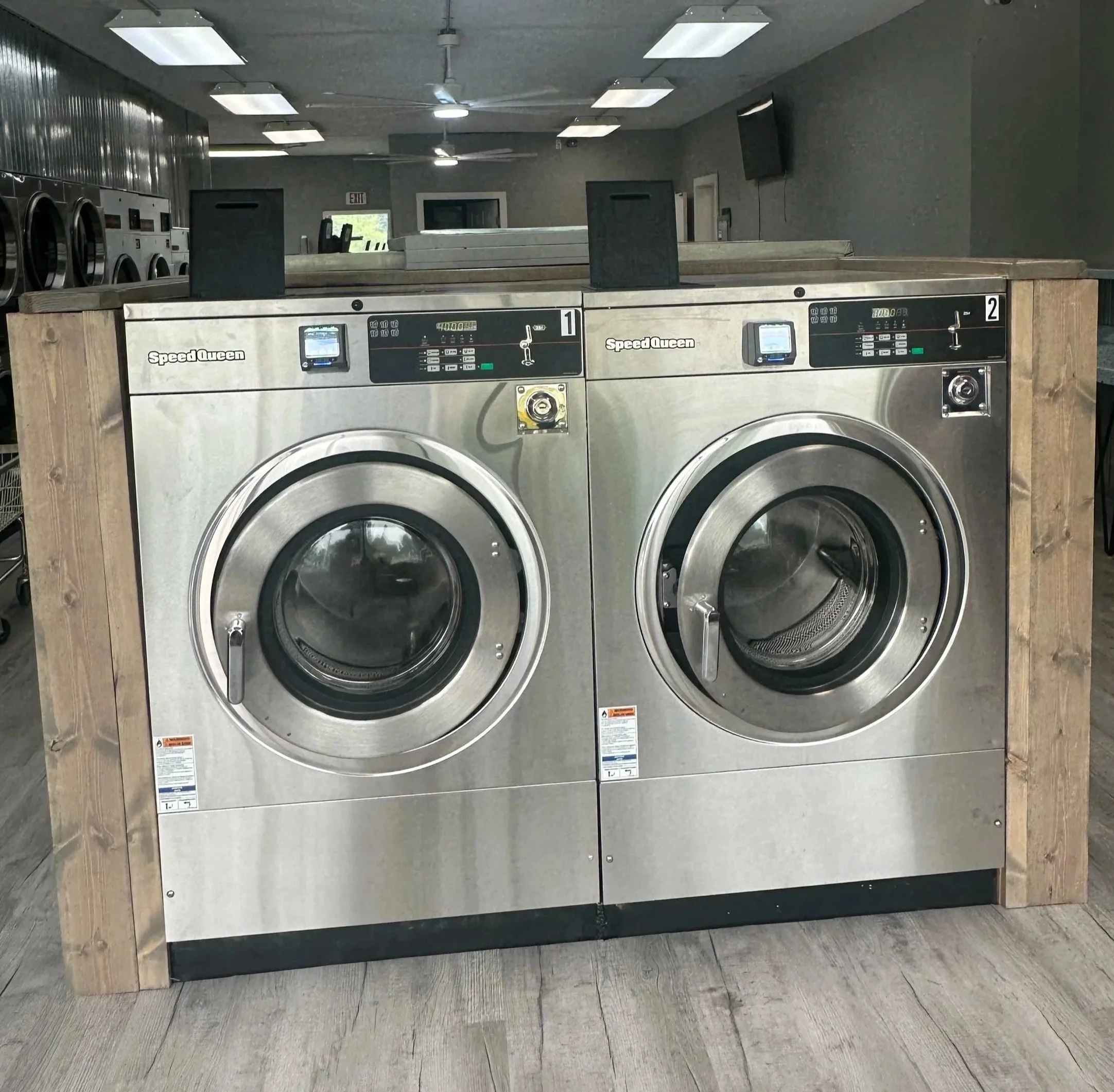 Interior of a laundromat with multiple industrial washing machines, metal folding tables, and laundry baskets. Recycling bin and yellow walls are visible.