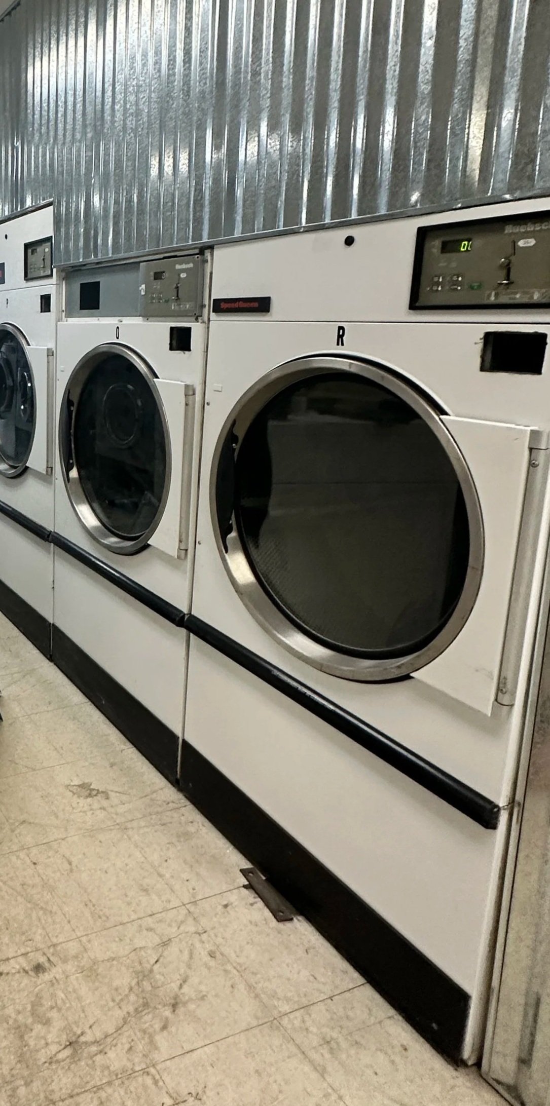 Row of stainless steel front-loading Speed Queen washing machines in a laundromat with visible control panels and glass doors.