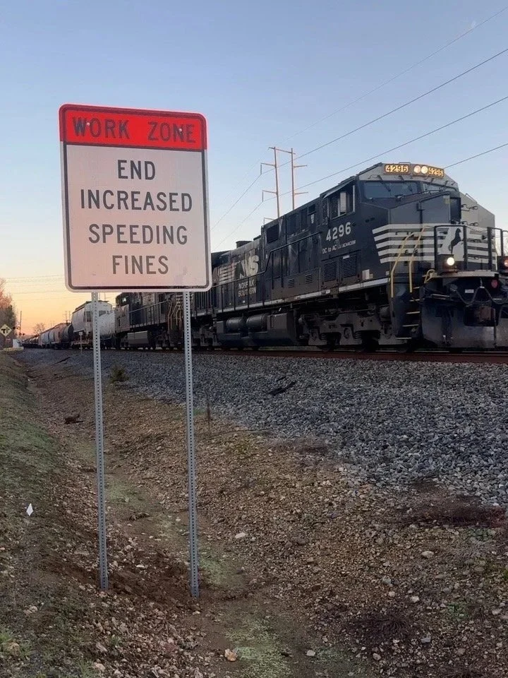 Railroad tracks with a passing train and a sign that reads 'Work Zone, End Increased Speeding Fines' against a backdrop of a partly cloudy sky.