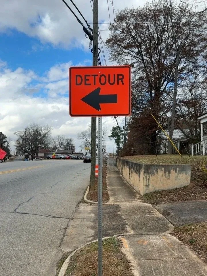 An orange detour sign with a black arrow pointing left, mounted on a pole along a suburban sidewalk with trees and houses in the background.