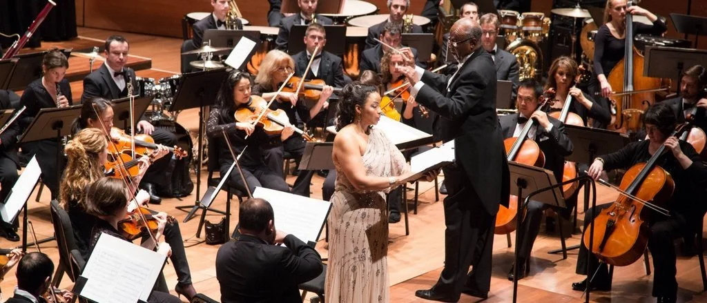 A symphony orchestra performing with a female soloist, Heather Hill, on stage. The conductor stands in front of the musicians, leading the performance. String and wind instruments are visible, and the musicians are wearing formal attire.