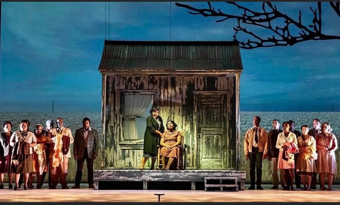 Theater stage scene with actors in front of a weathered wooden house. Two central actors are positioned under a spotlight, while a group of actors stands on either side of the house, dressed in period clothing. The backdrop features a blue sky and tree silhouette.