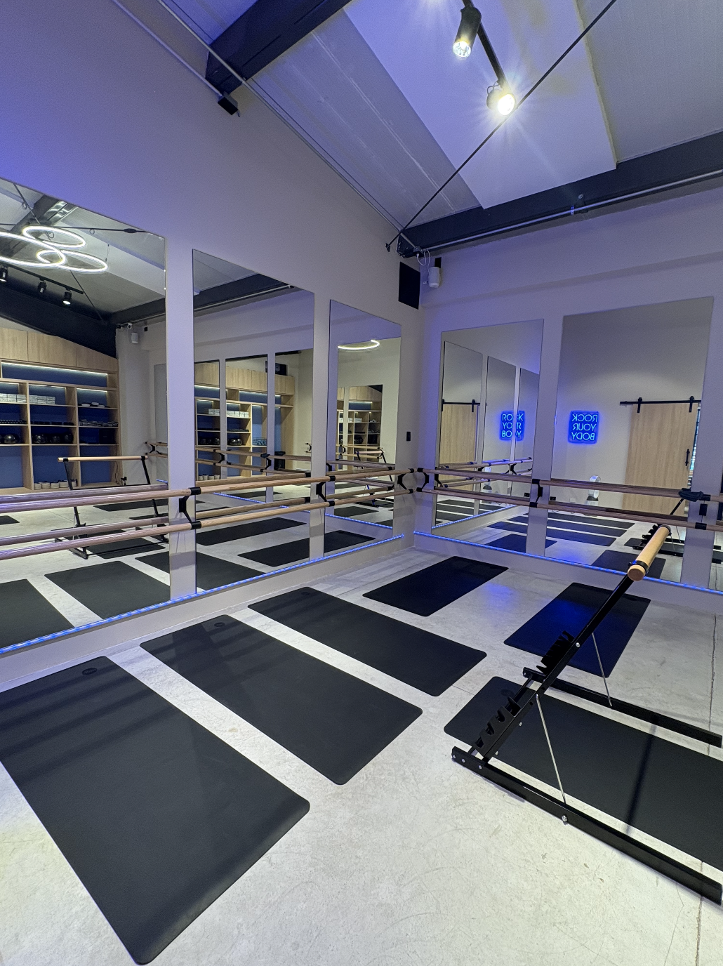 Estudio de yoga y barre con tapetes negros, espejos en las paredes, barras de ballet, y luces LED azules, decorado con techo moderno.