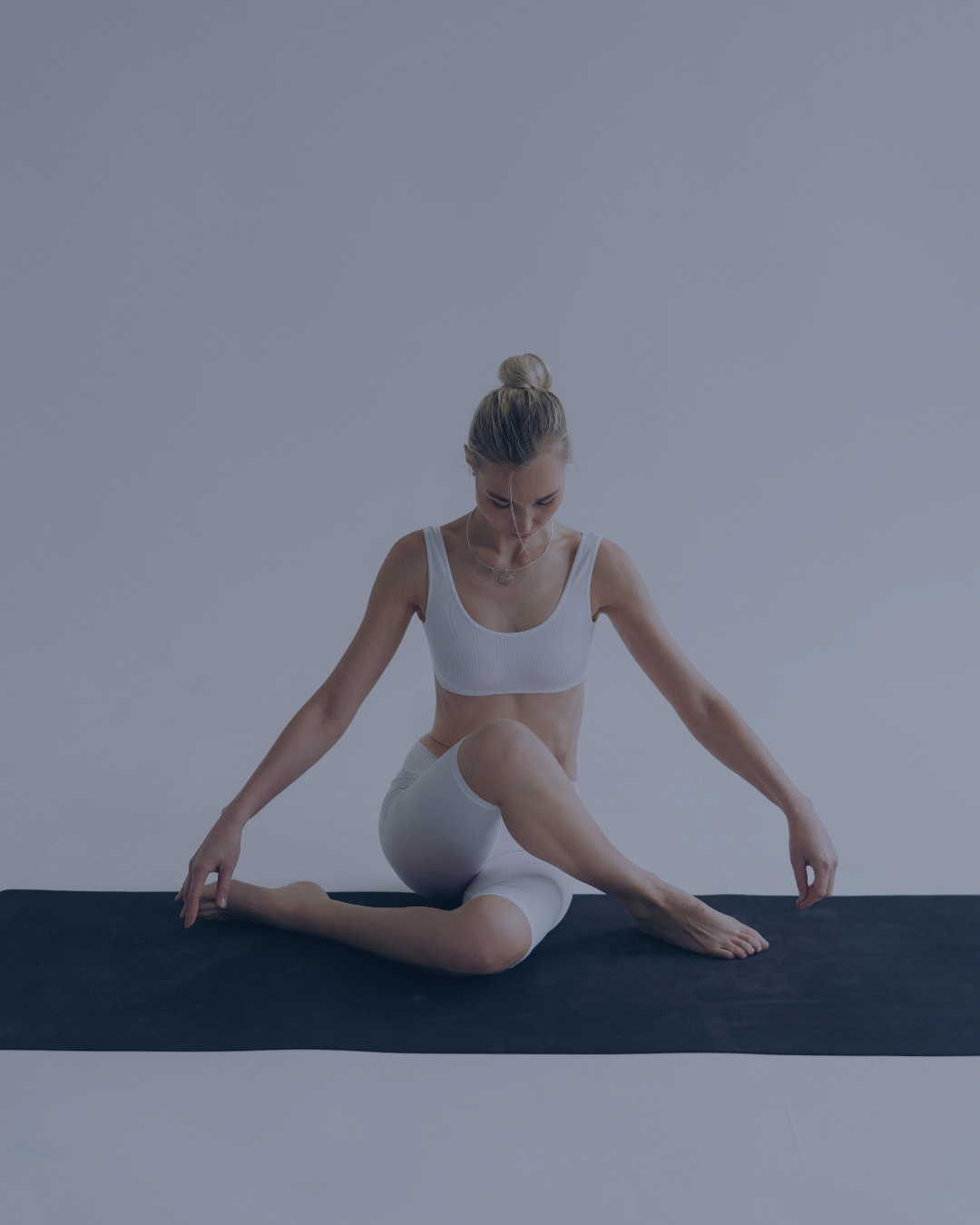 Mujer haciendo yoga en una colchoneta negra, con ropa blanca, en un fondo gris.