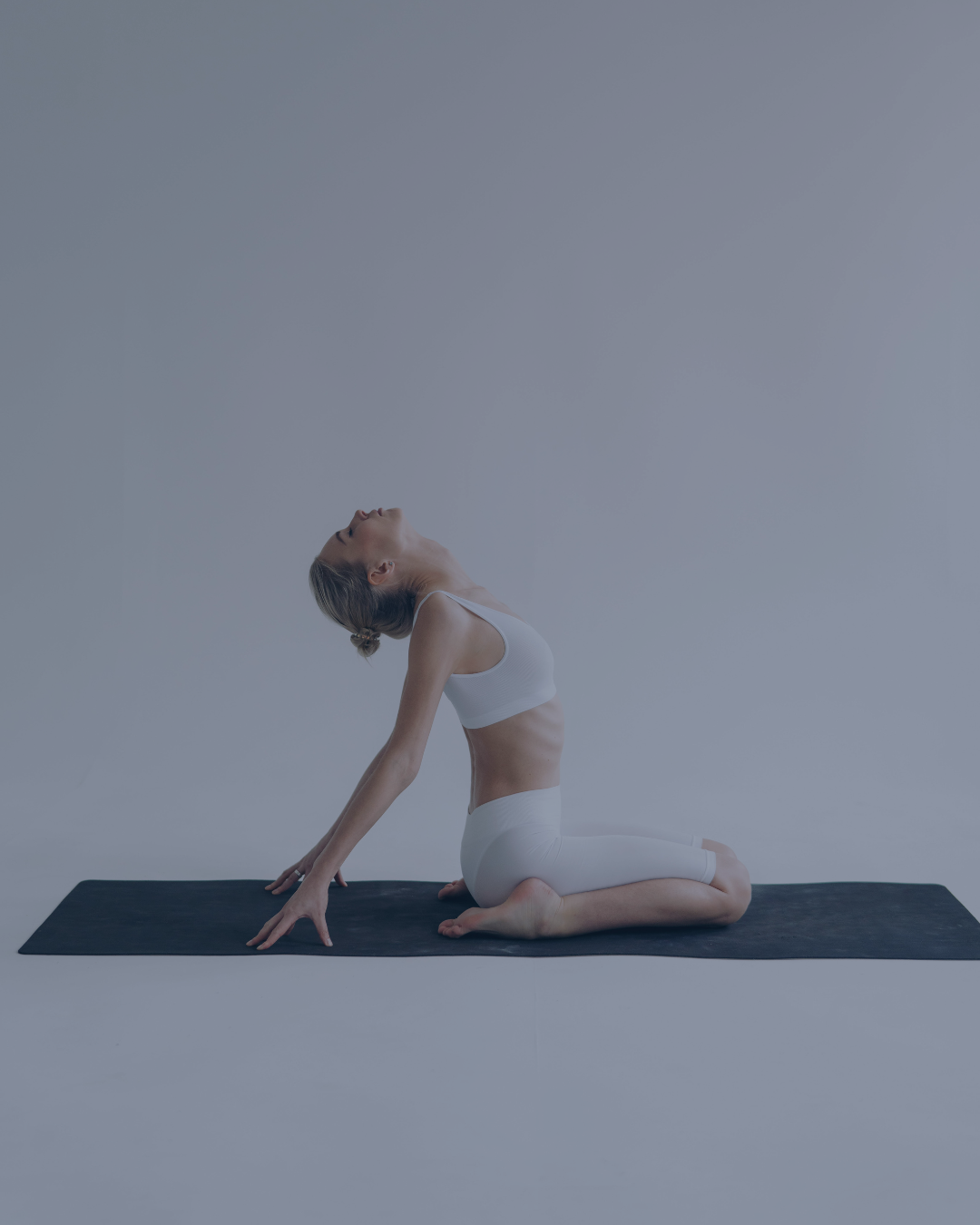 Mujer practicando yoga en una posición de arrodillada con el pecho levantado y la cabeza inclinada hacia atrás, sobre una esterilla negra en un ambiente minimalista.