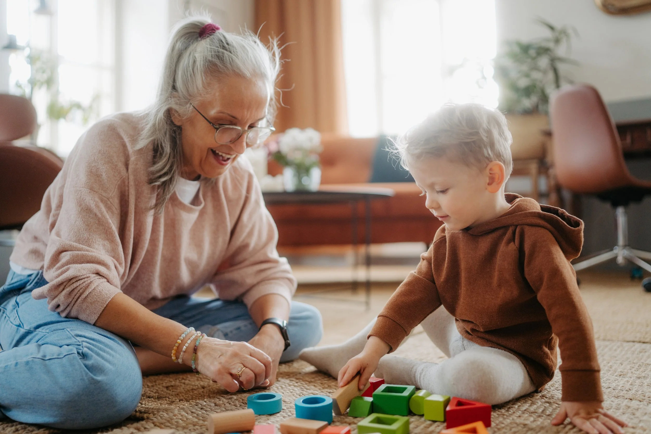 An elderly woman and a young child playing with colorful building blocks on a carpeted floor in a bright living room.