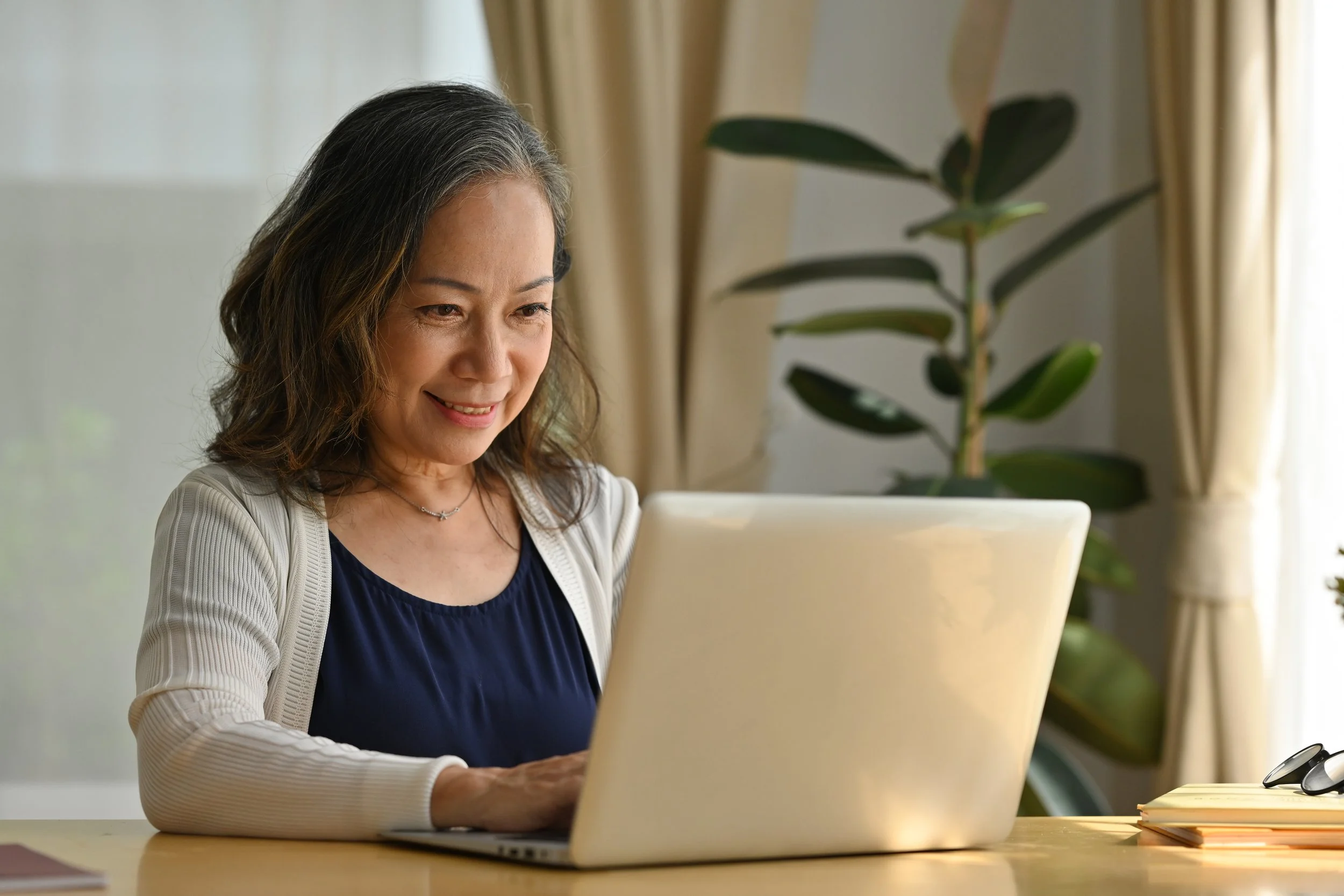 A middle-aged woman with dark hair and a smile working on a laptop at home with a large potted plant and set of glasses on the desk.