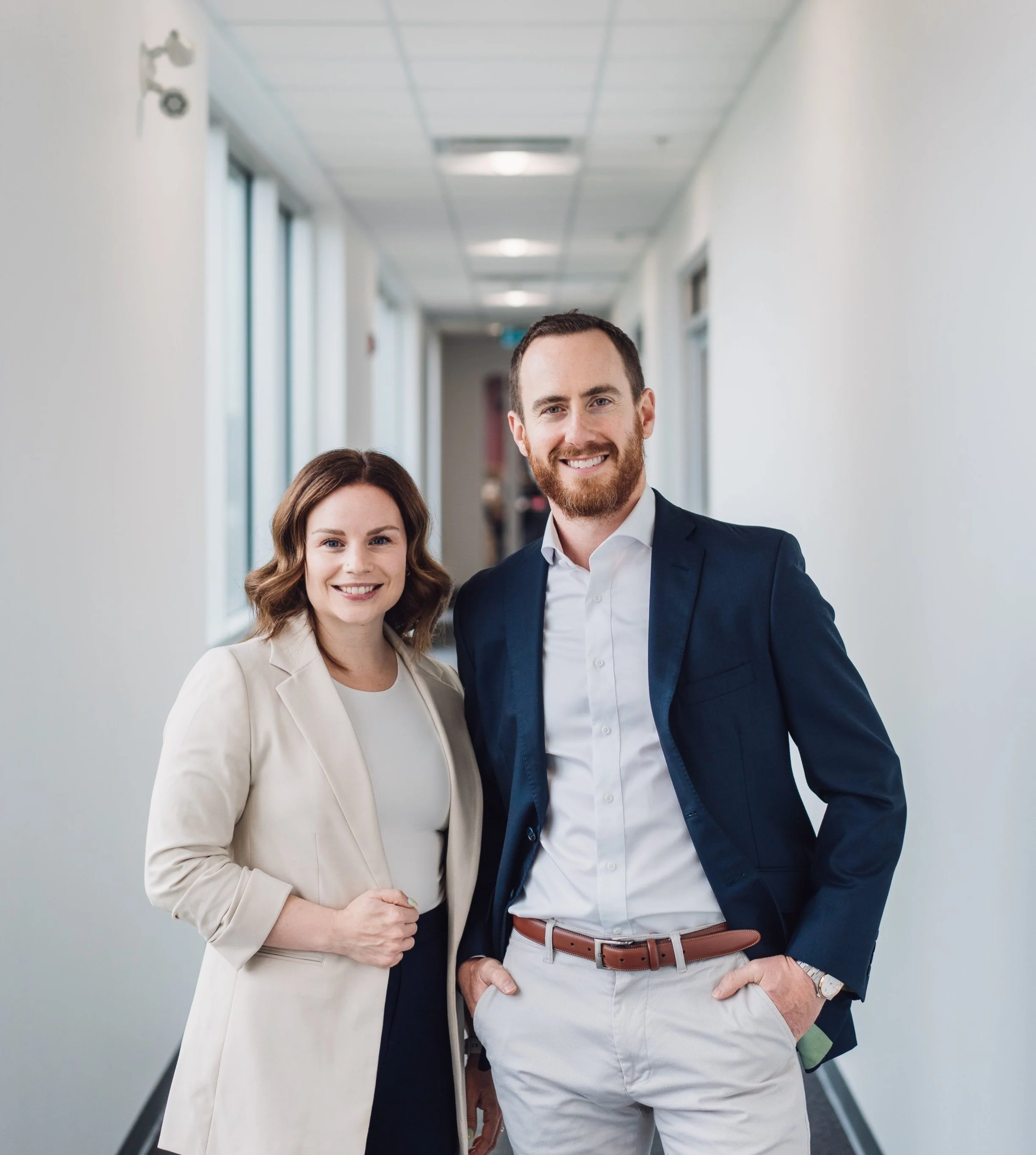 Hayley and Noah standing together in a hallway, smiling at the camera, dressed in professional attire.