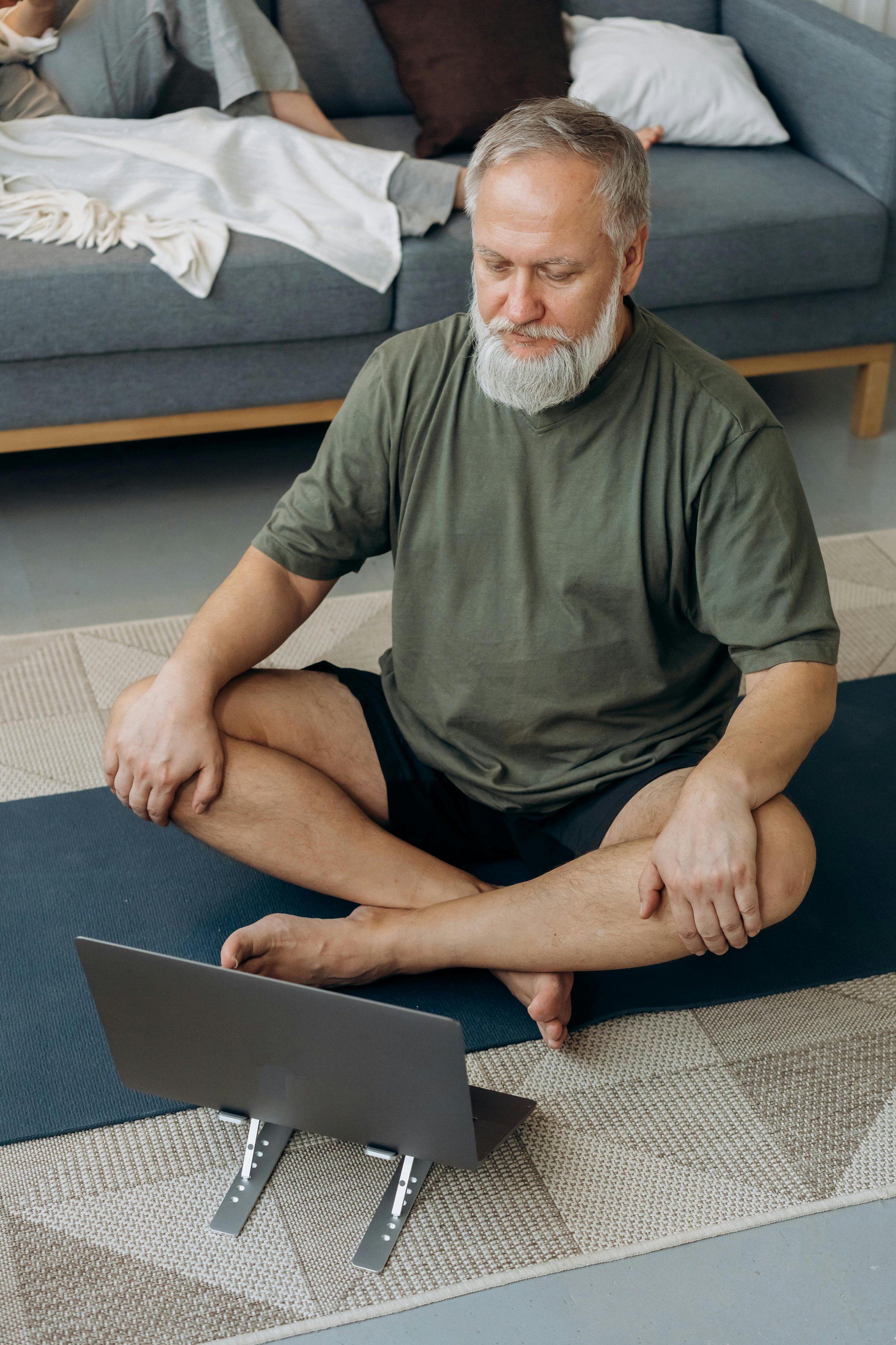 A man practicing yoga indoors sitting on a mat in a cross-legged position, looking at a laptop in front of him.