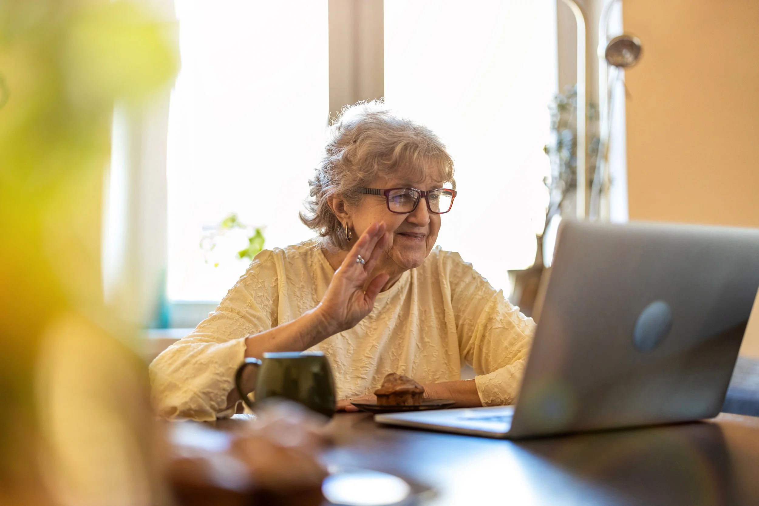 An elderly woman with glasses smiling and waving on a video call while sitting at a table with a laptop and mug, in front of her, near a window.