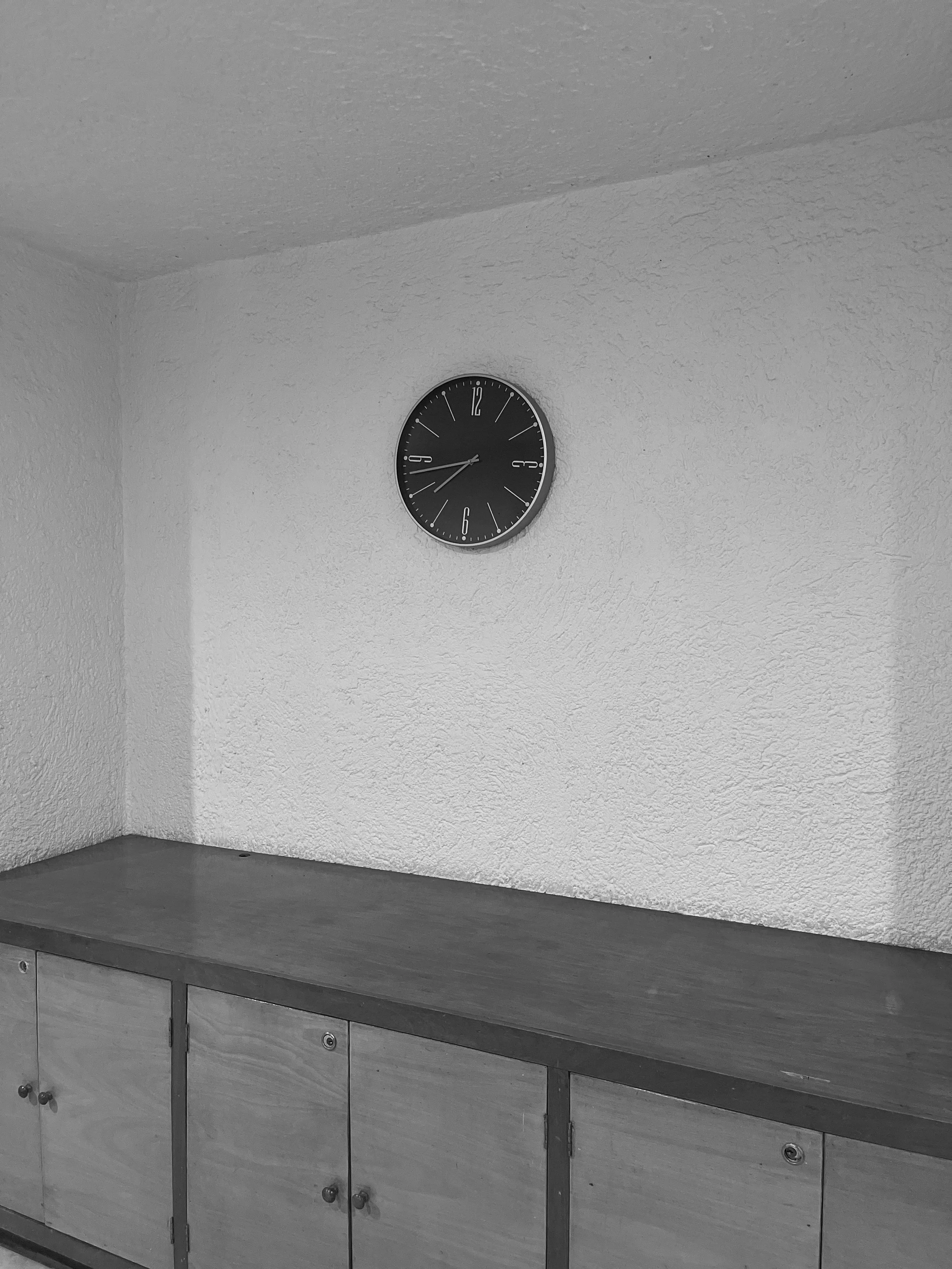 Black and white photo of a wall clock on a textured wall above a wooden cabinet.