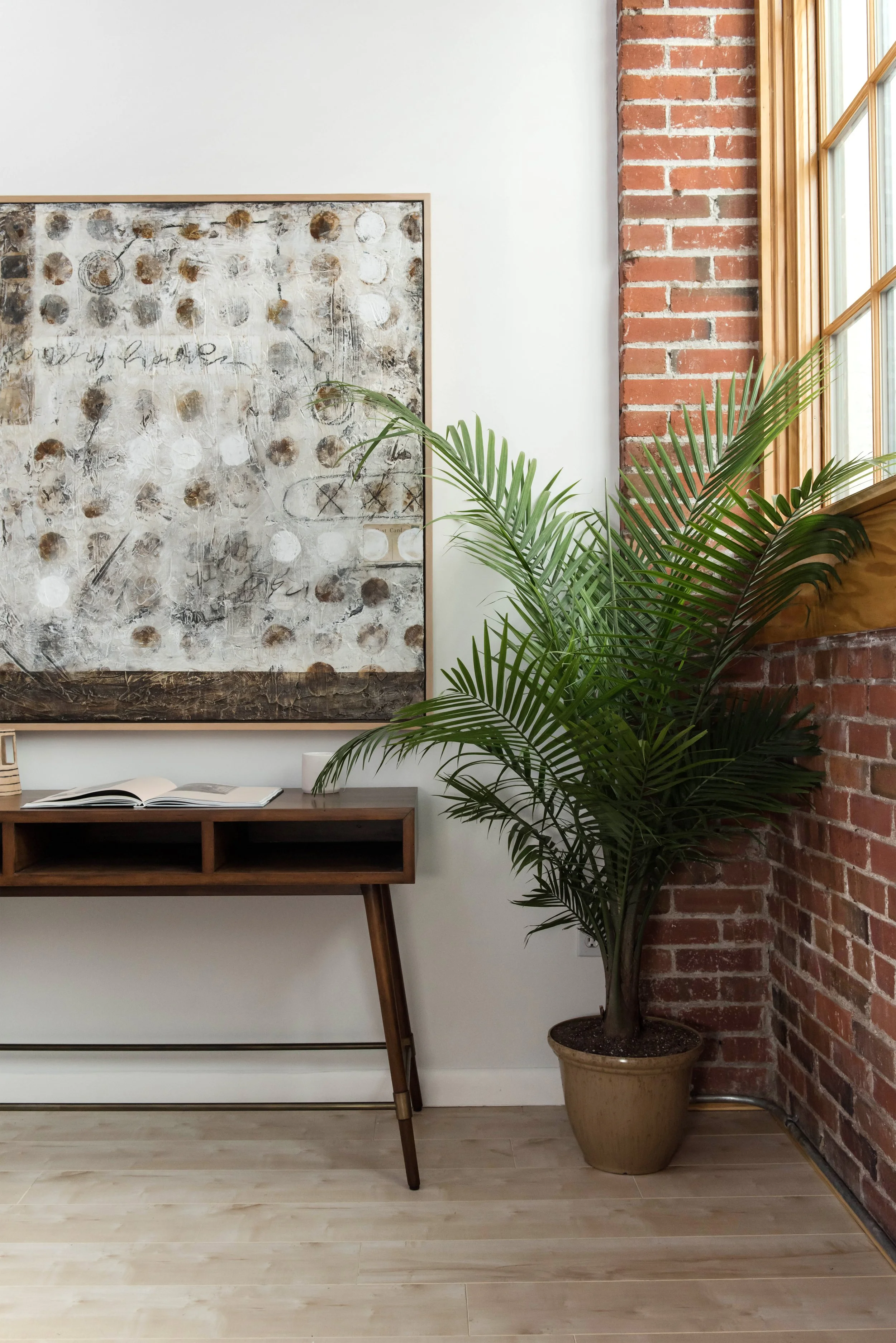 Interior of a room with a brick and white wall, a large abstract painting on the wall, a wooden table with an open book, and a potted palm plant near a window.