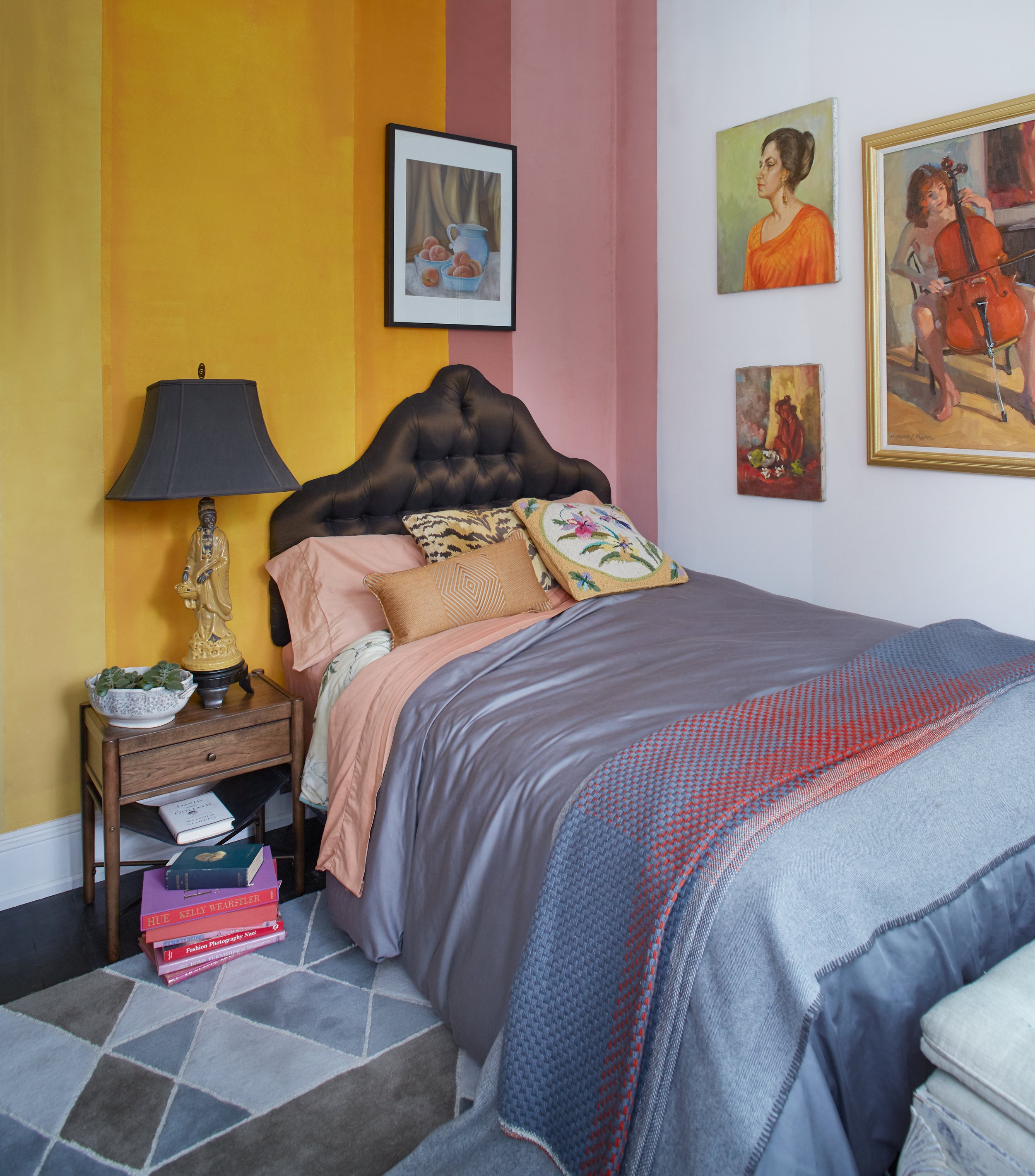Cozy bedroom with colorful bedding, striped yellow and pink walls, and a gallery of paintings. Features a tufted headboard, bedside table with a lamp and statue, and a stack of books on the floor. Residential Design, Renovation, Restoration, Interior