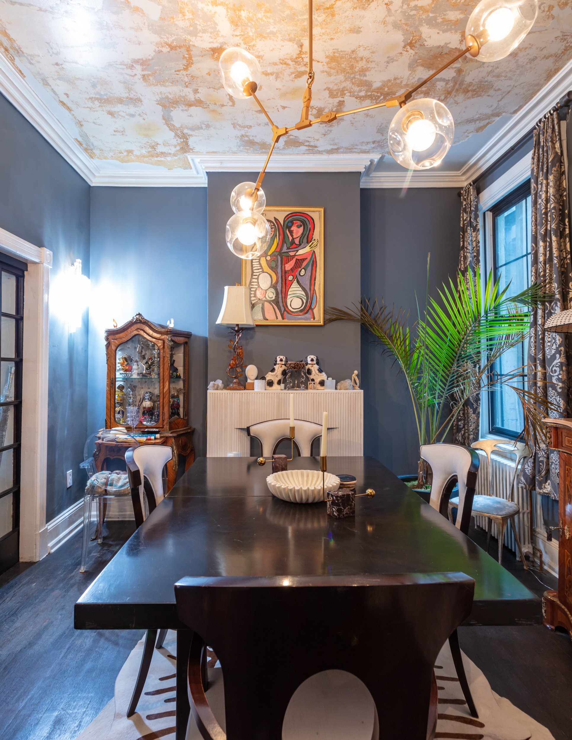 Eclectic dining room with a dark wood table, modern light fixture with spherical bulbs, abstract wall art, and decorative items on a mantle. The walls are blue and there is a mix of vintage and contemporary decor. Residential Design, Renovation, Rest
