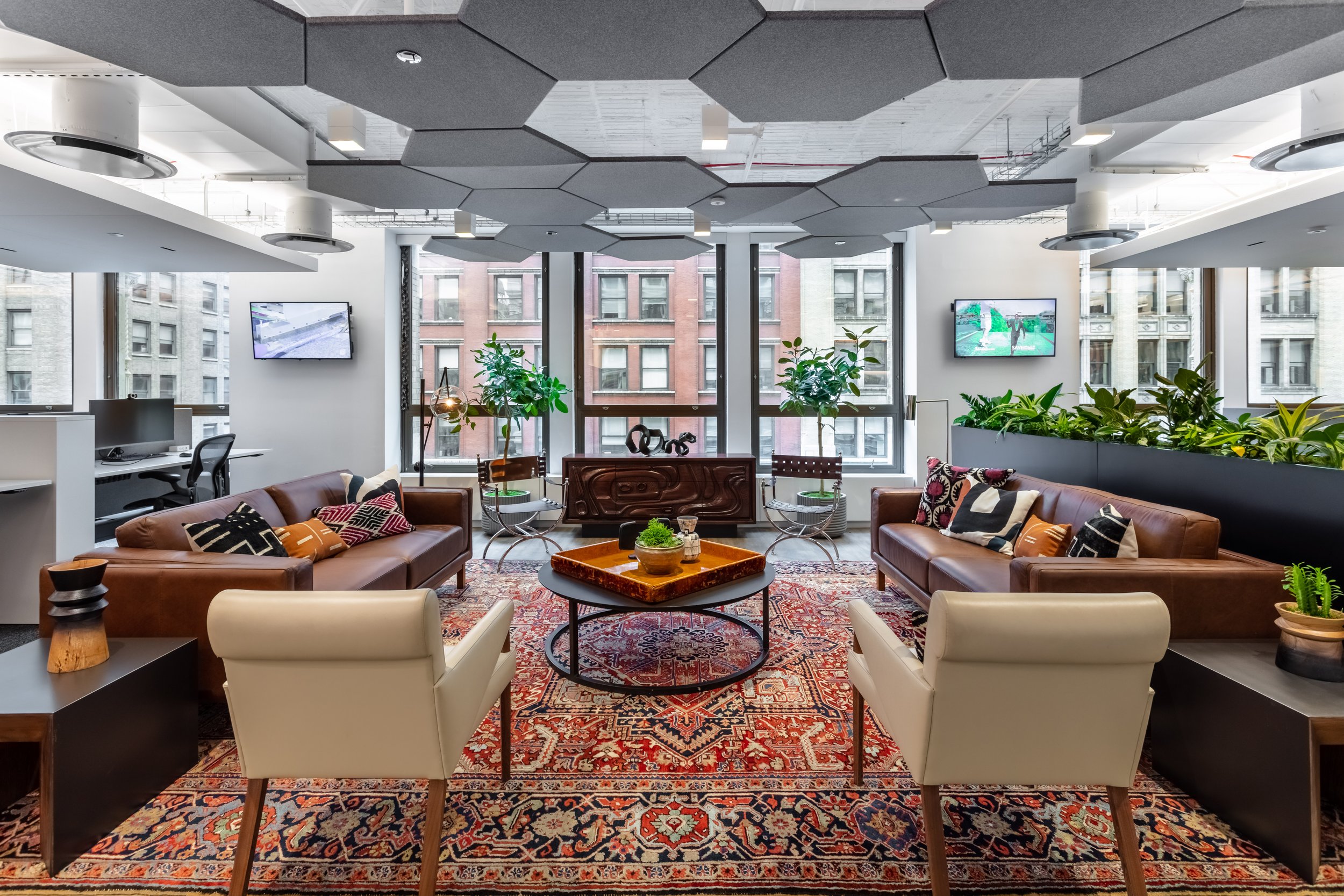Modern office lounge area with leather sofas, armchairs, patterned rug, potted plants, and large windows. The ceiling features geometric acoustic panels, and there are multiple flat-screen TVs on the walls. Interior Furnishings, Decor, Accessories an
