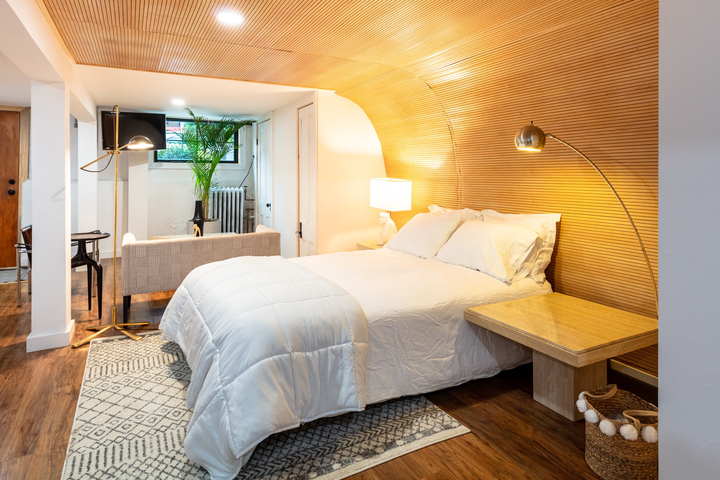 Cozy bedroom with a modern design, featuring a wooden accent wall and ceiling, double bed with white linens, bedside lamps, a patterned rug, and a small sofa facing the foot of the bed. A potted plant and radiator are near a window in the background.