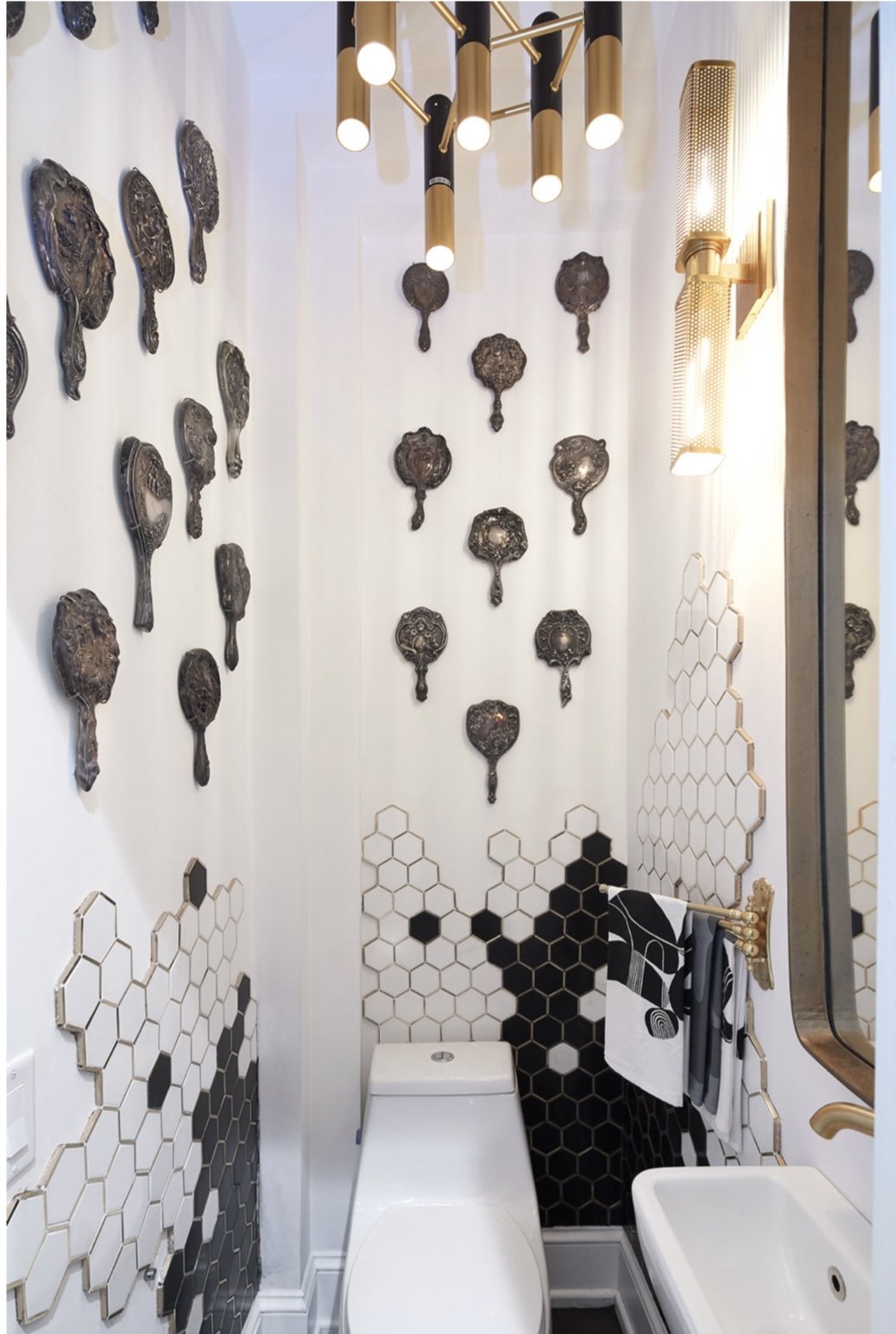 A small bathroom with hexagonal black and white tile design, antique hand mirror wall art, modern lighting fixtures, a toilet, and a towel rack with patterned towels. Residential Design, Renovation, Restoration, Interior Design and Decor, Art Curatio