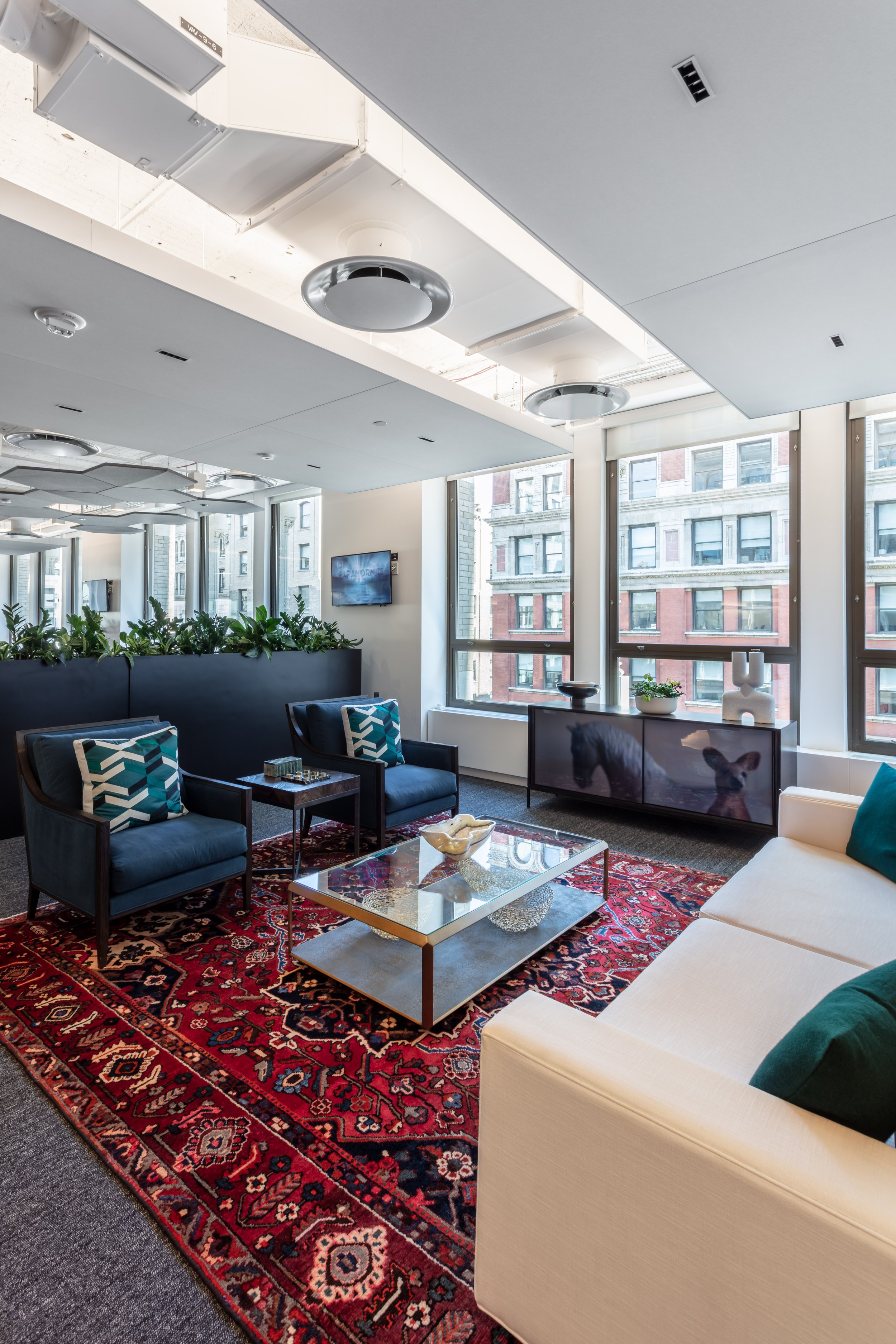 Modern office lounge with white sofa, glass coffee table on red patterned rug, two navy armchairs, and large windows showing cityscape. Interior Furnishings, Decor, Accessories and Art, Design, Procurement, Hope Misterek