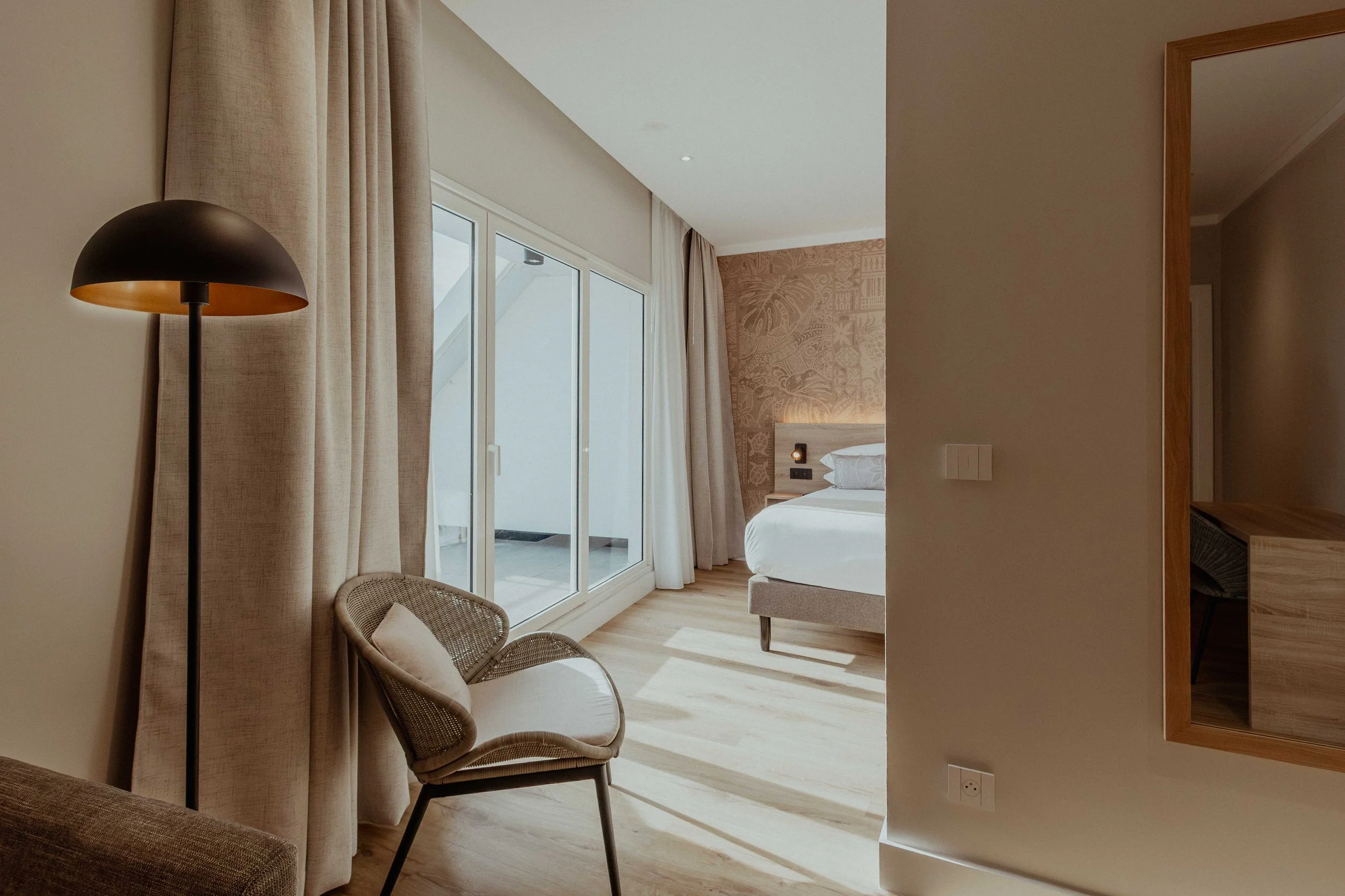 Chambre élégante avec un lit blanc, une peau de tête décorative et une grande vitre donnant sur un balcon, avec un fauteuil en osier et un lampadaire noir.