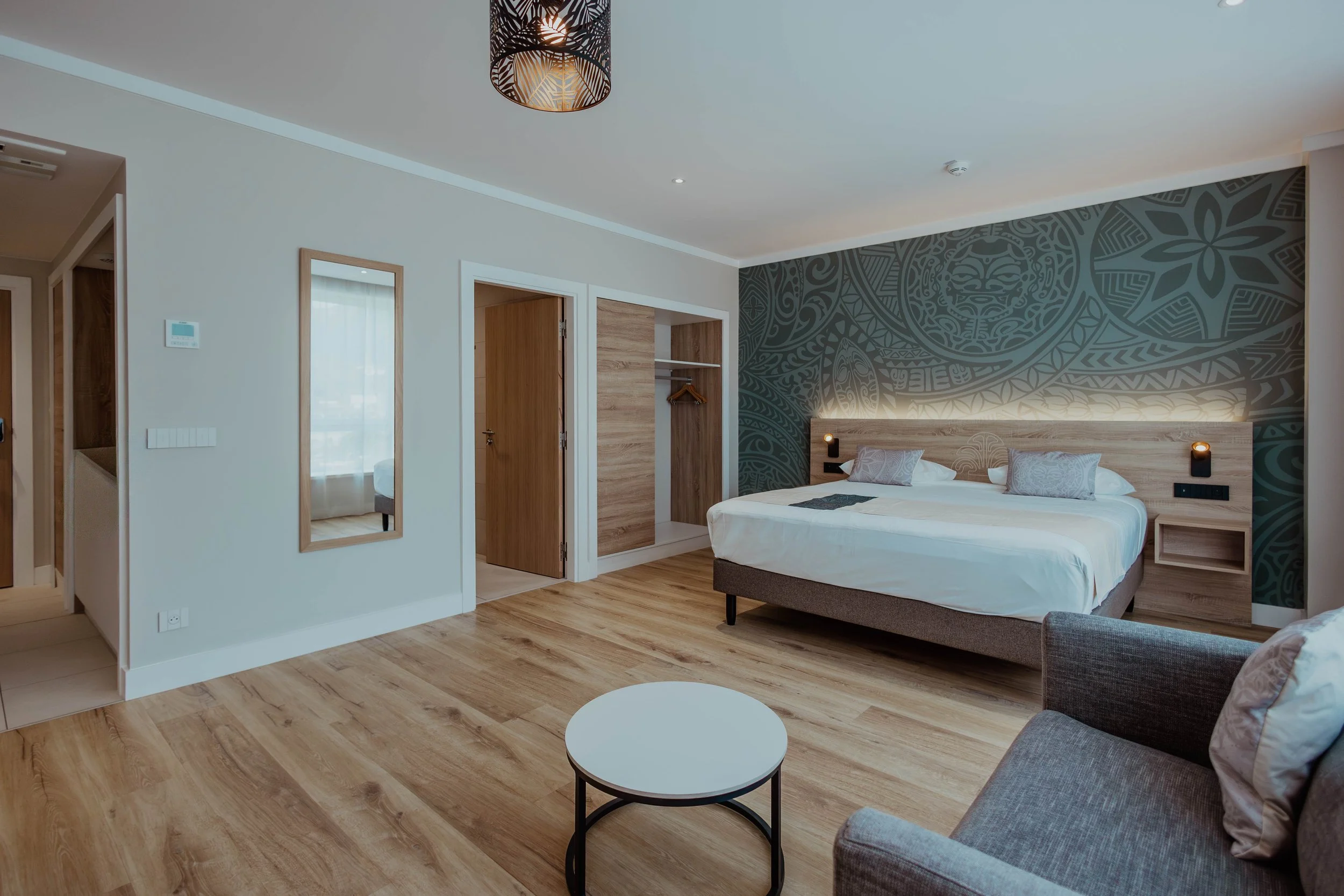 Chambre d'hôtel avec un grand lit, deux coussins, une tête de lit en bois clair, un mur de décoration avec un motif tribal, un coin salon avec un canapé gris et une petite table ronde blanche.