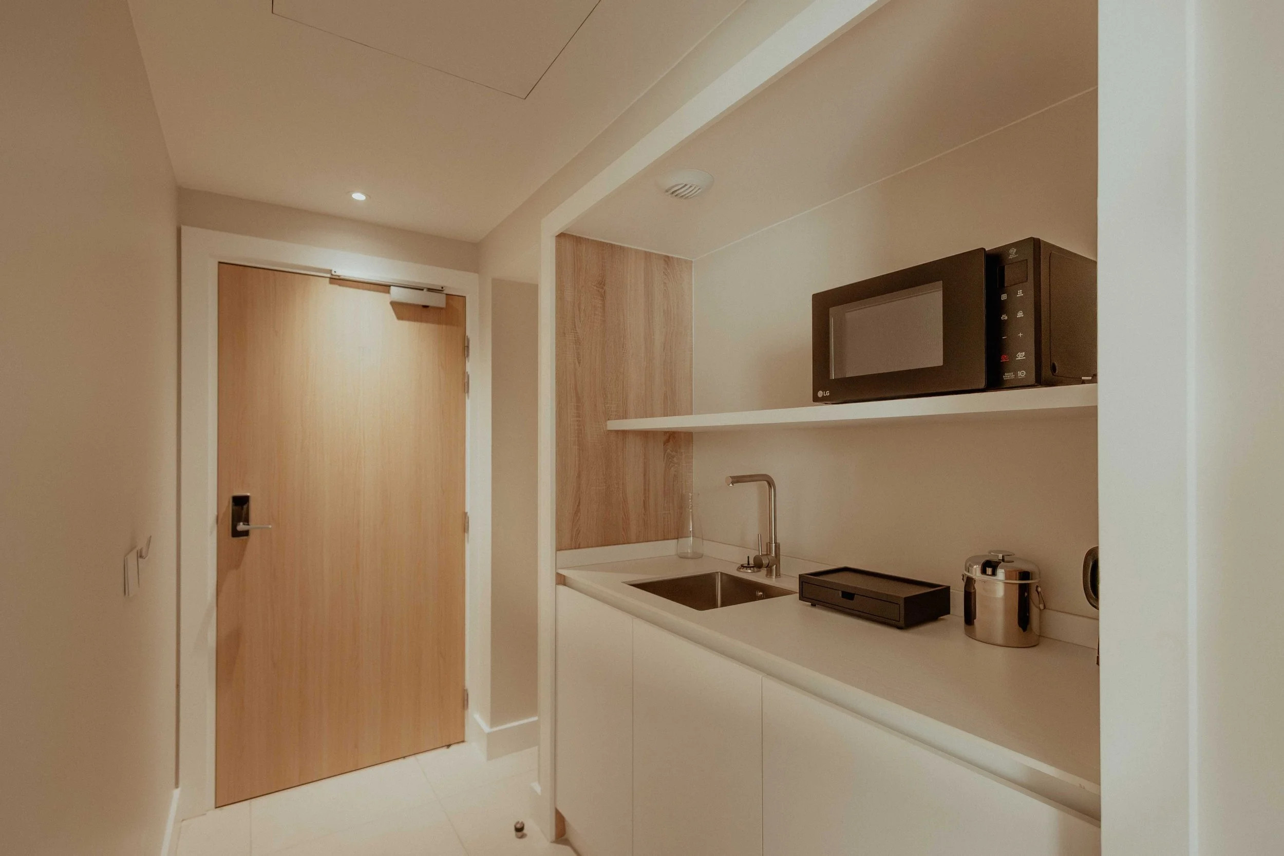 Petite cuisine moderne avec un évier, un micro-ondes, une plaque électrique, une bouilloire électrique et un porte-serviette, dans un espace minimaliste avec murs blancs et porte en bois.