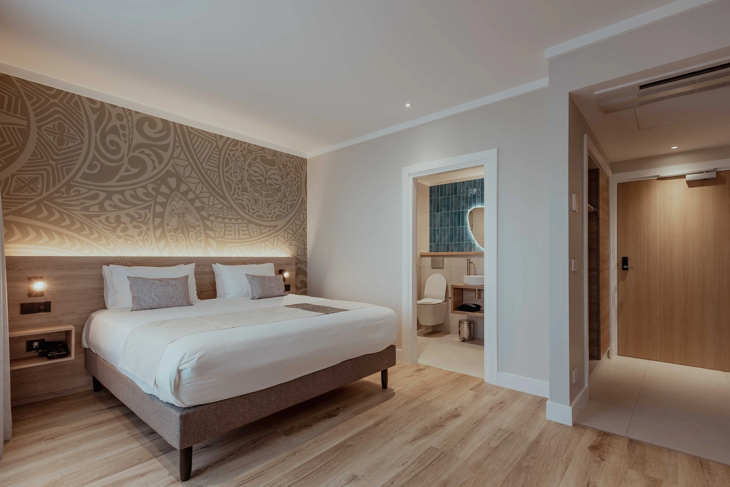 Chambre d'hôtel moderne avec lit double, mur décoratif derrière le lit, petite salle de bain avec toilettes et lavabo, porte en bois.