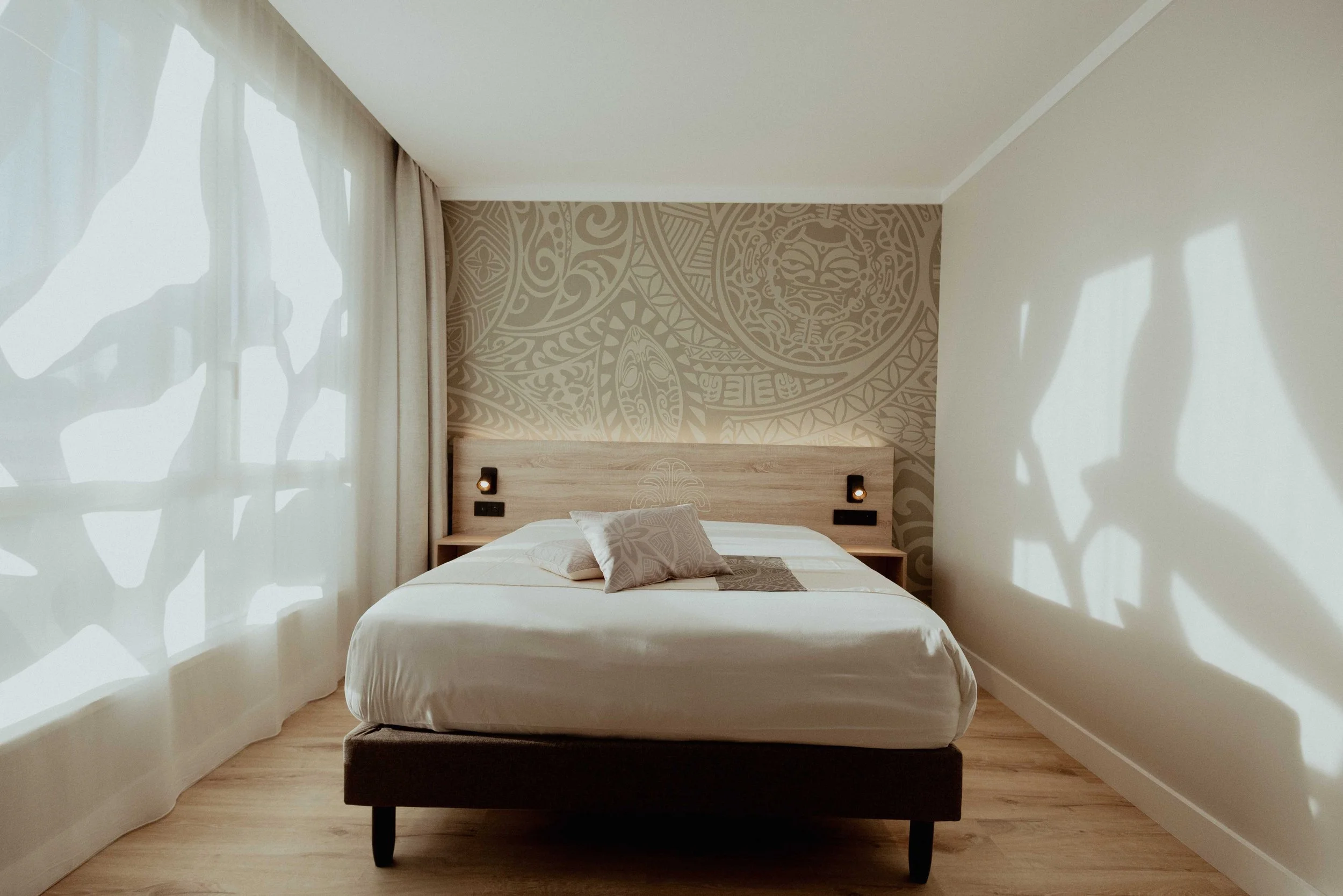 Chambre avec un lit, deux coussins, une tête de lit en bois, décor mural beige à motifs Polynésie, rideaux blancs laissant passer la lumière