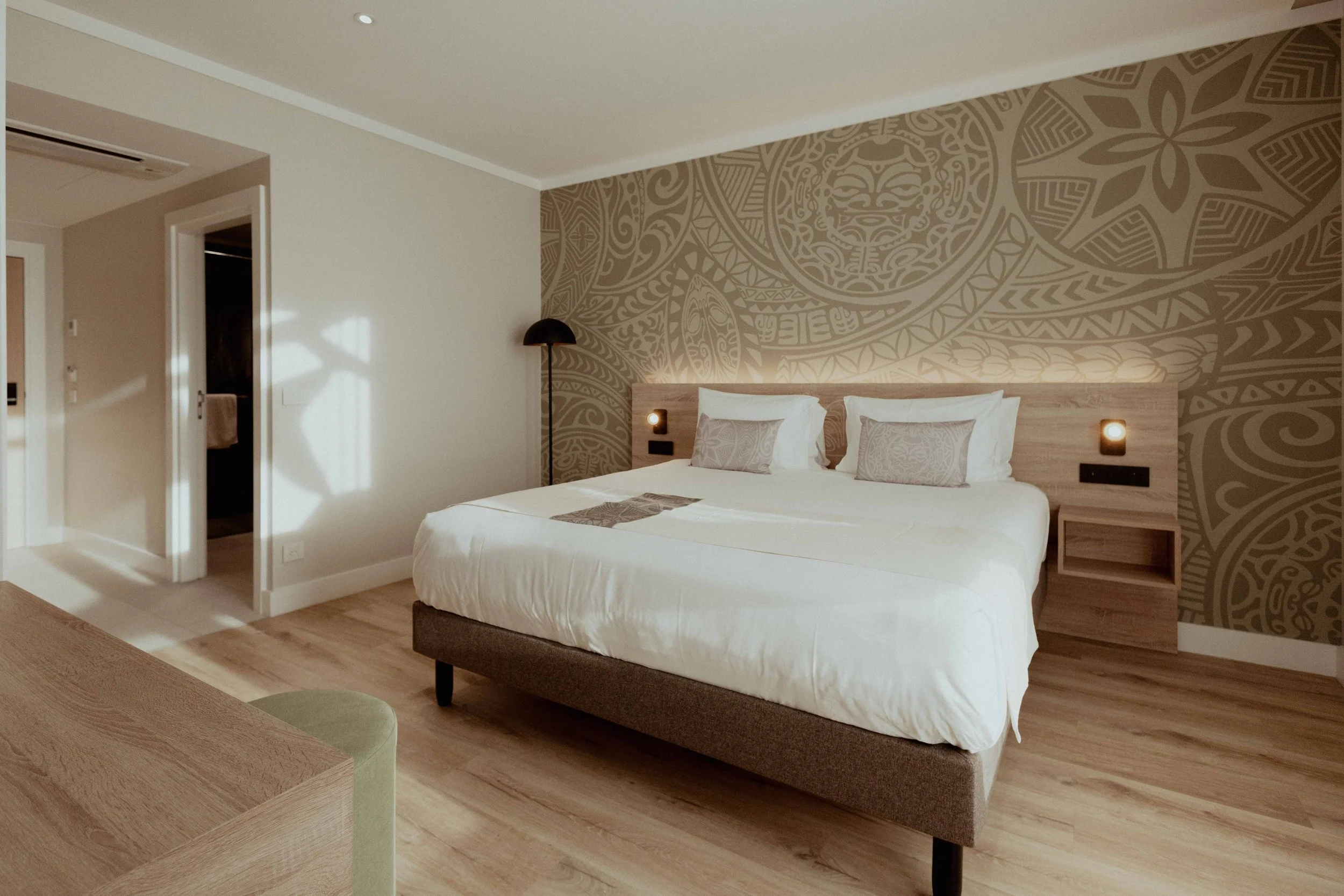 Chambre d'hôtel moderne avec lit king-size, mur en papier peint avec motifs tribaux, lampe de chevet, et plancher en bois.