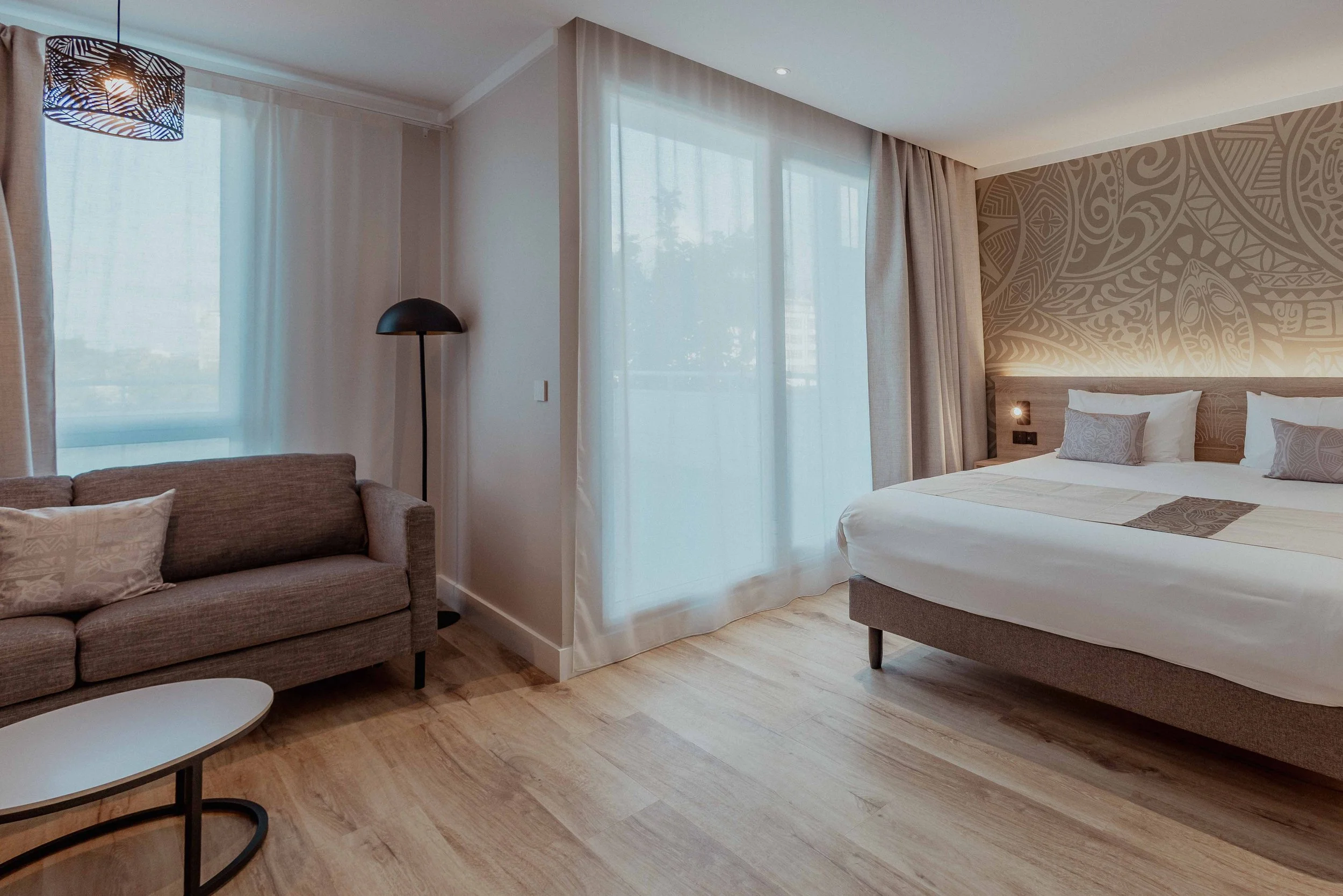 Chambre d'hôtel moderne avec grand lit, décoration sobre, canapé en tissu, lampadaire noir, fenêtre avec rideaux légers, papier peint avec motifs abstraits derrière le lit, sol en bois clair.