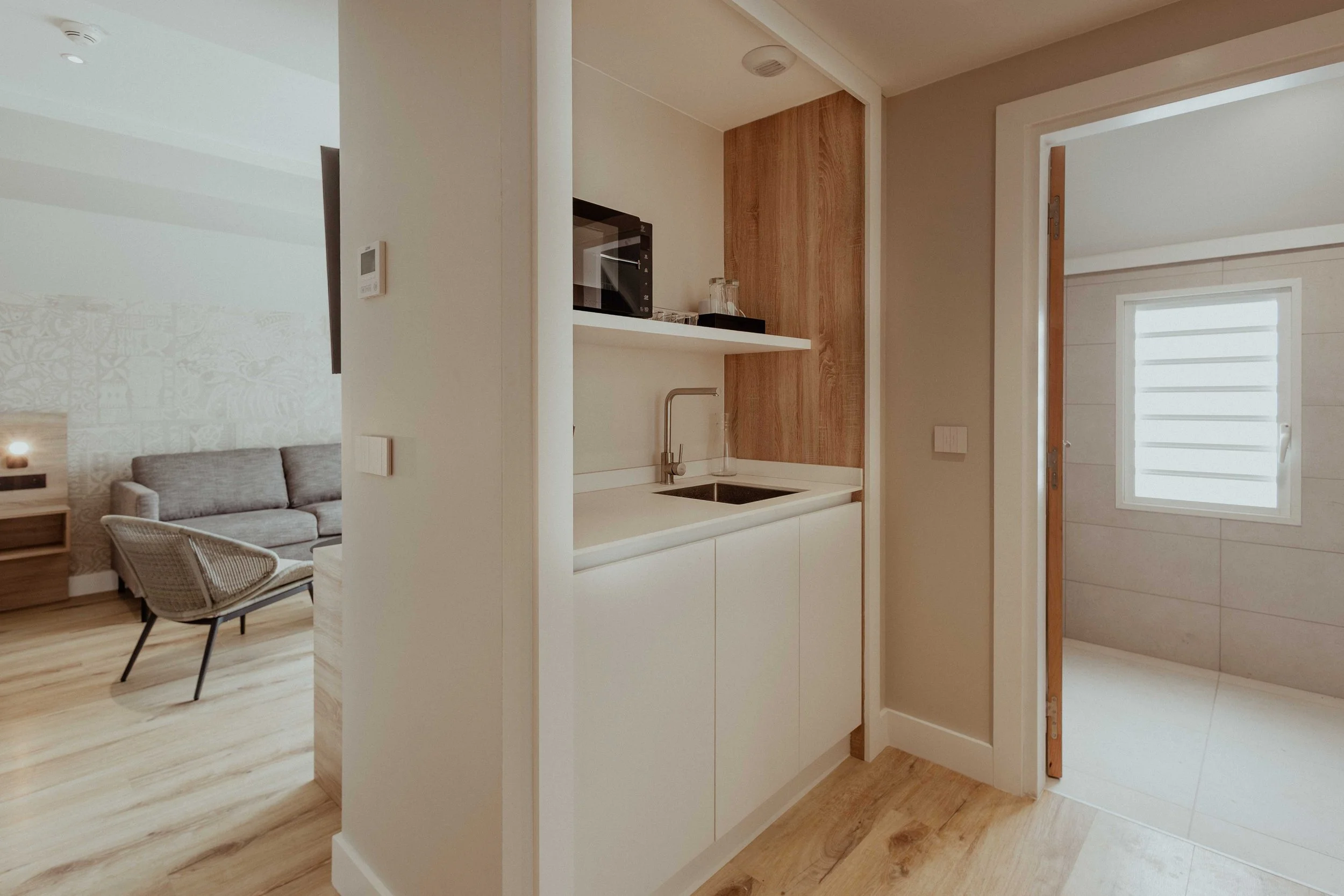 Coin cuisine moderne avec un évier, un micro-ondes sur une étagère, une fenêtre au fond et une pièce de vie avec un canapé gris et une chaise en rotin.