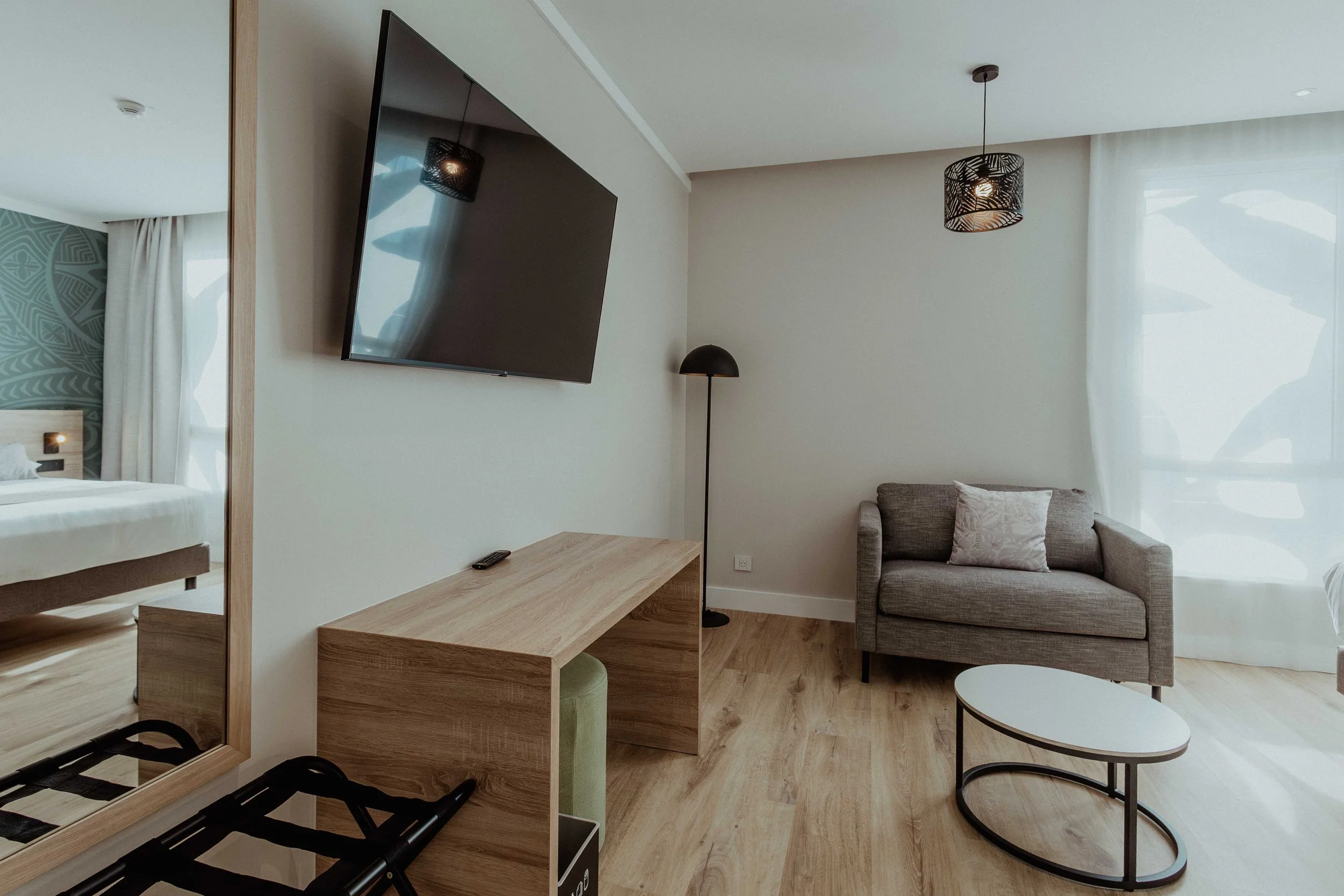 Salon avec canapé gris, table basse ovale, lampadaire noir, télévision murale, fenêtre avec rideaux transparents, chambre visible au fond à gauche.