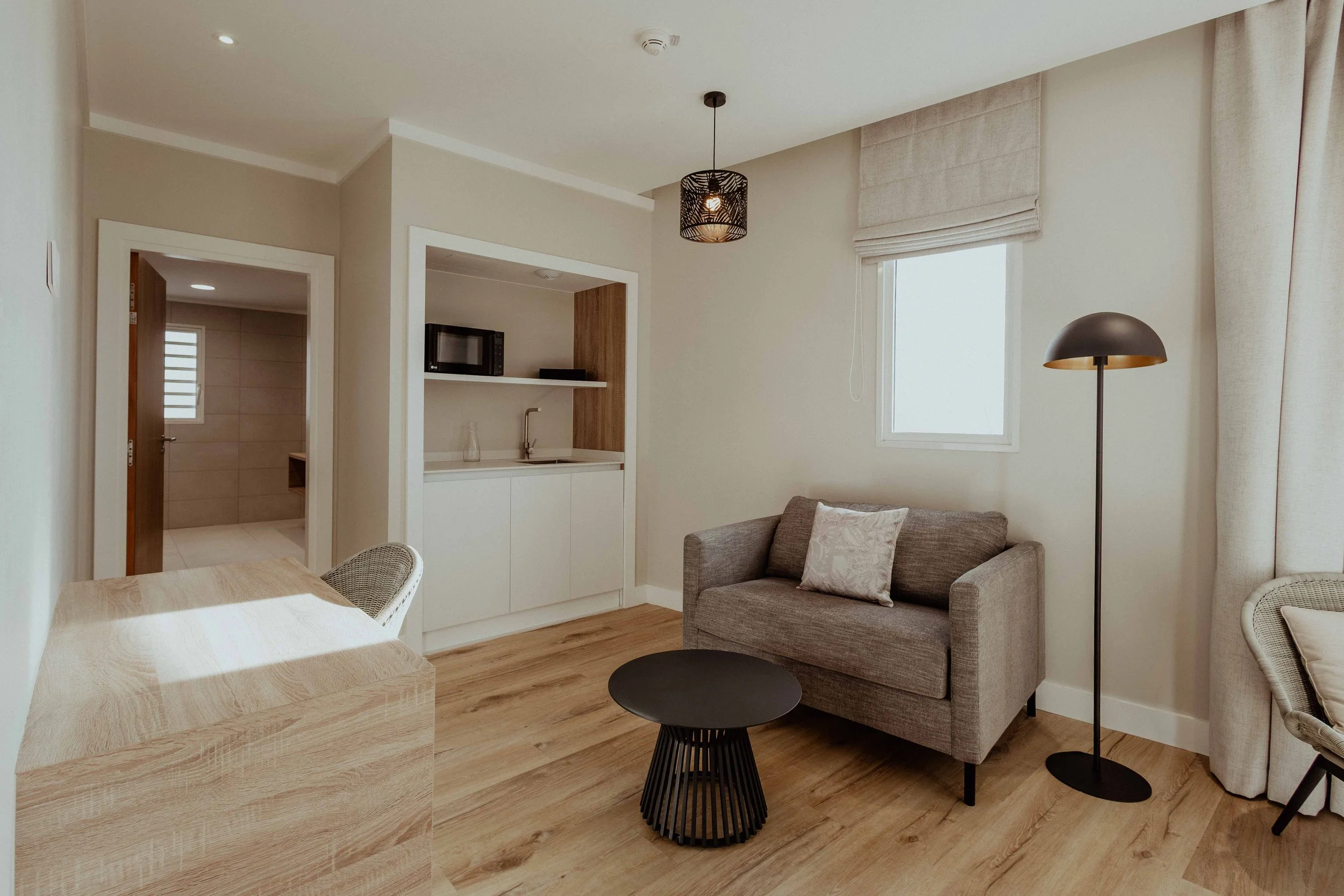 Salon avec canapé gris, lampe sur pied noire, table basse ronde noire, fenêtre avec rideaux beige, cuisine ouverte avec micro-ondes, évier et étagère, porte menant à une salle de bain.