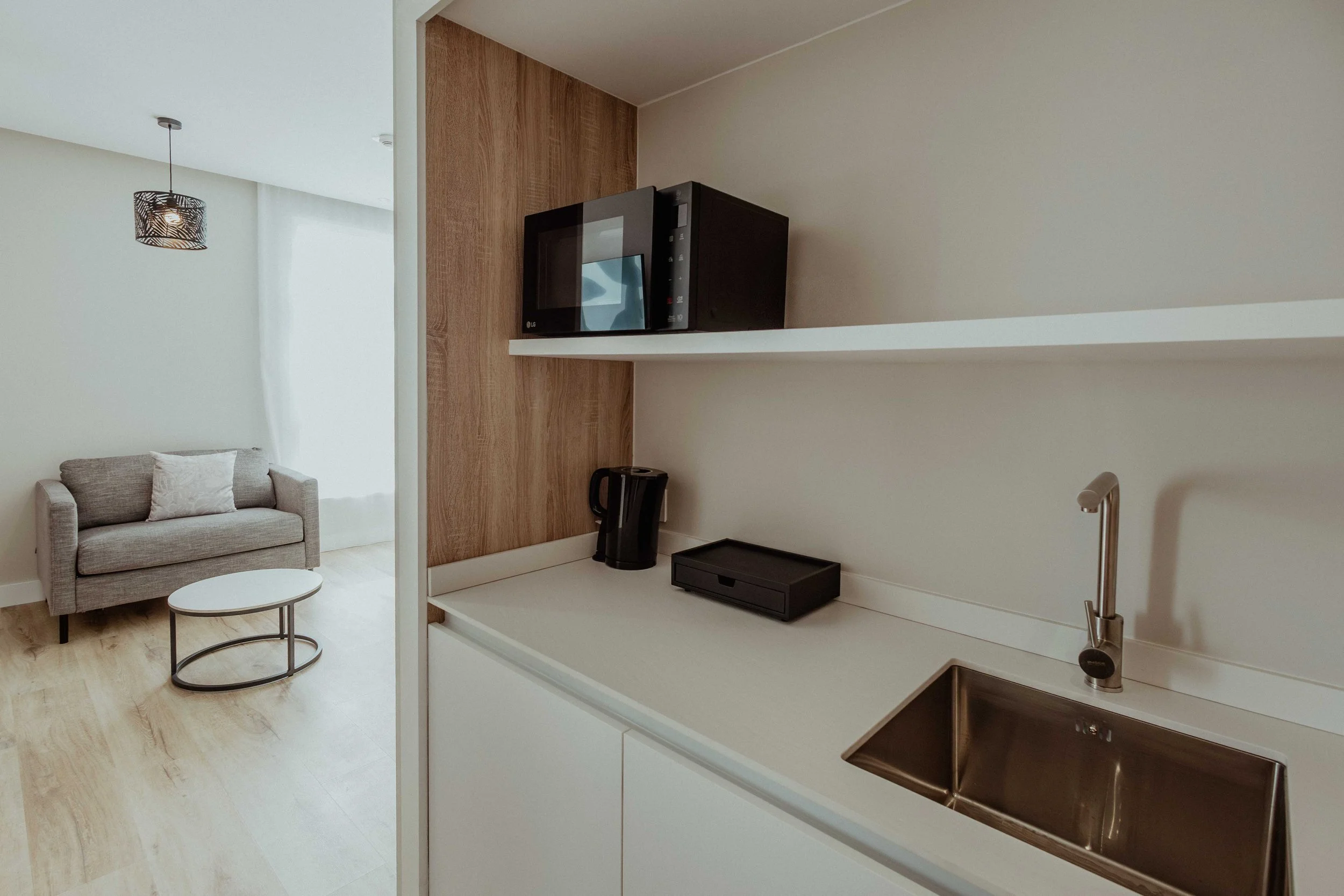 Coin cuisine avec évier, micro-ondes, théière, étagère, salon avec canapé et table dans un appartement moderne.