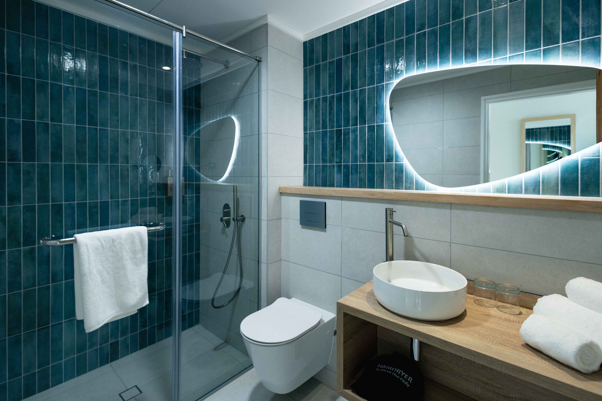 Salle de bain moderne avec douche en verre, miroir éclairé, lavabo blanc sur étagère en bois, et toilettes suspendues, murs en carrelage bleu et blanc.