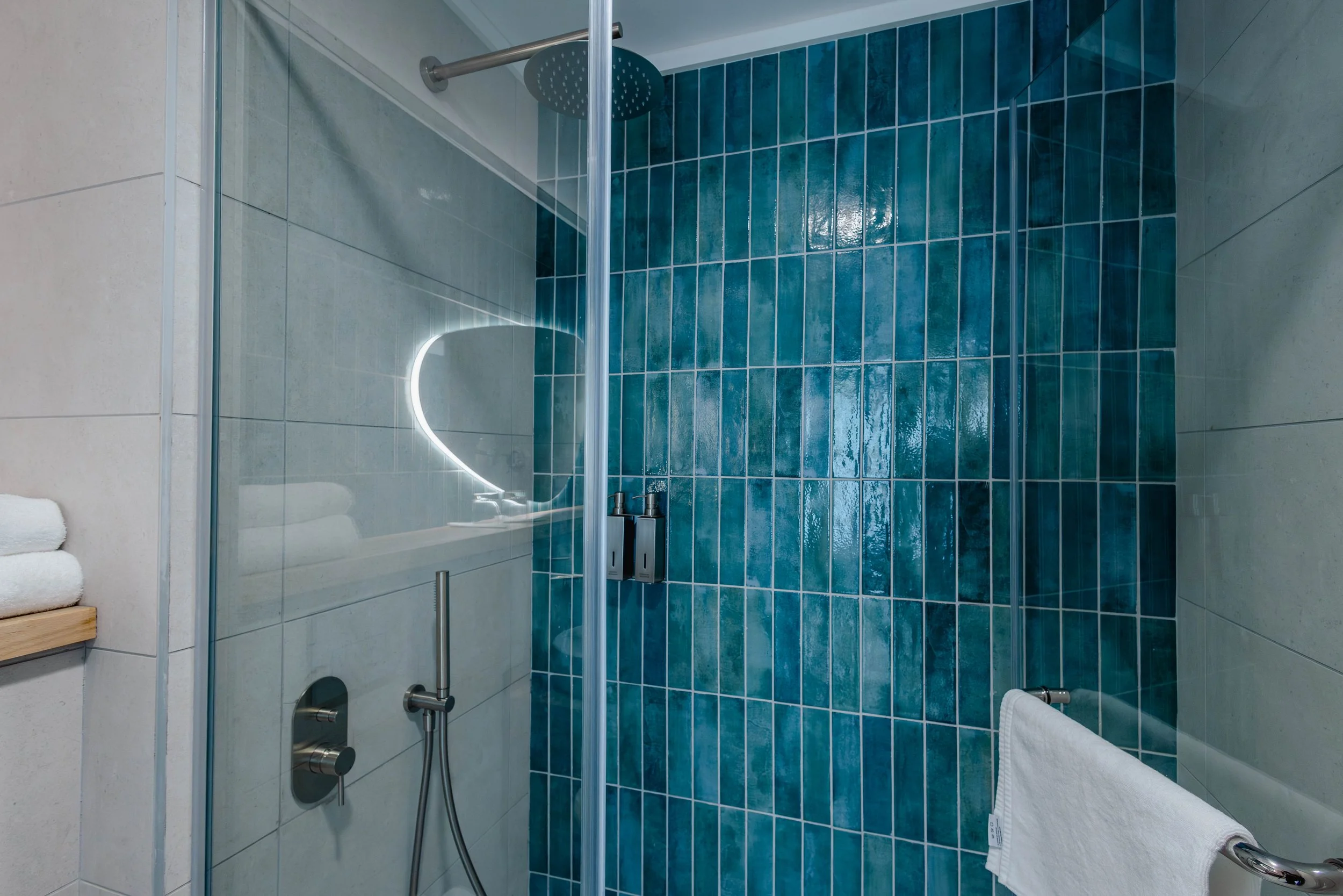 Salle de bain avec douche, carreaux bleus et blancs, miroir illuminé, serviette blanche sur barrière, étagères avec serviettes.