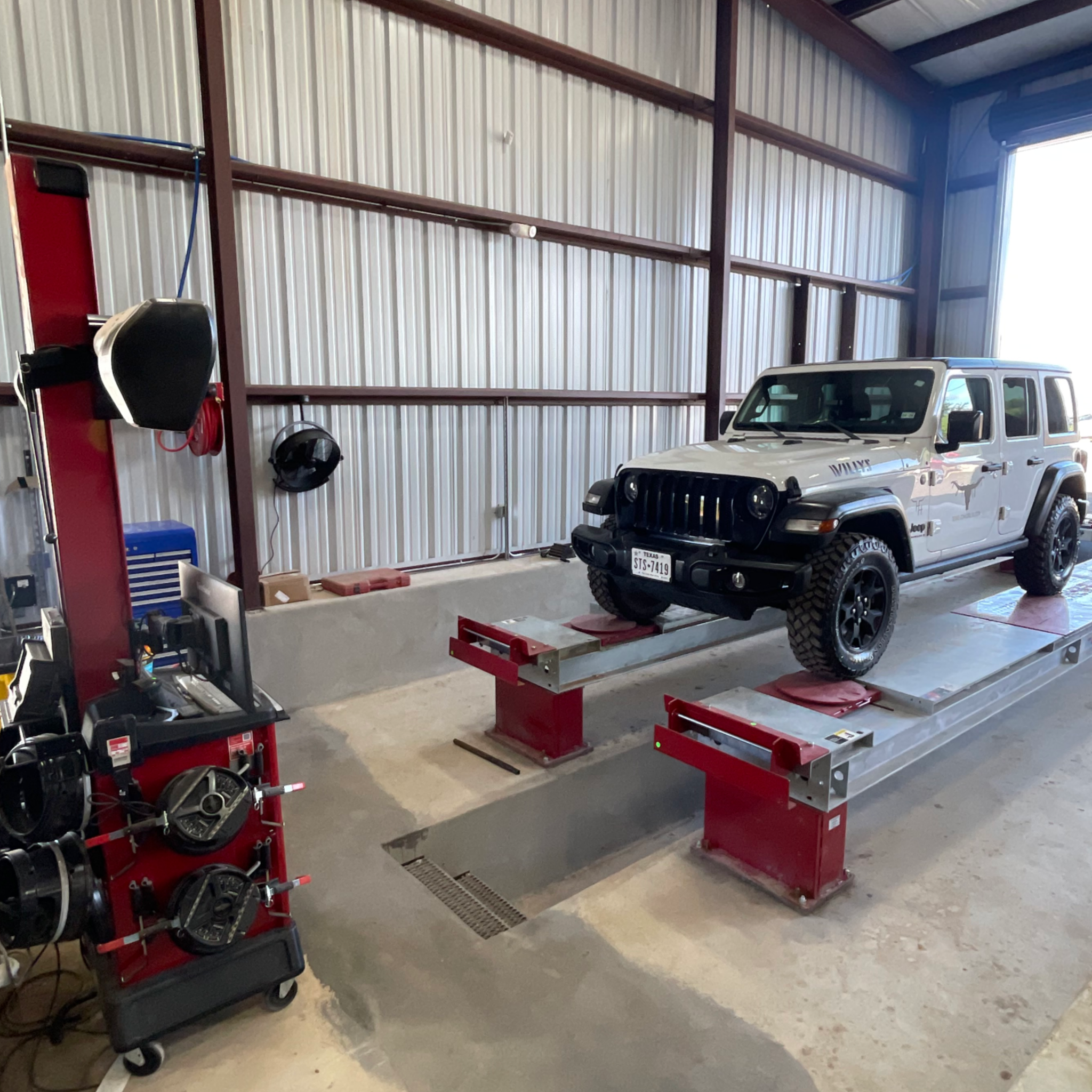 A white Jeep Wrangler on a hydraulic lift inside an automotive repair shop. The shop has a metal interior with tools and equipment nearby.