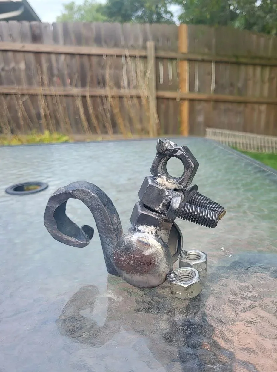 A sculpture made from metal scraps resembling a chameleon with a curled tail, placed on a glass table outdoors.