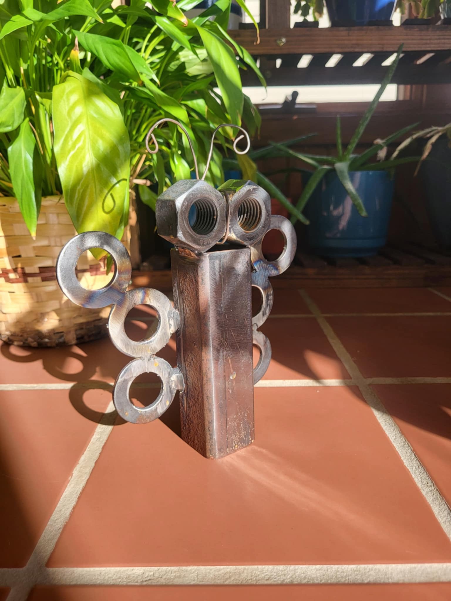A robot figure made from metal nuts, bolts, and washers, with wire antennae, standing on a tiled floor with potted plants and a wooden fence in the background.