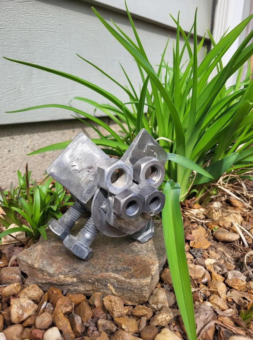 A metal mechanical part with multiple threaded holes, positioned among green plants and rocks outside a building.