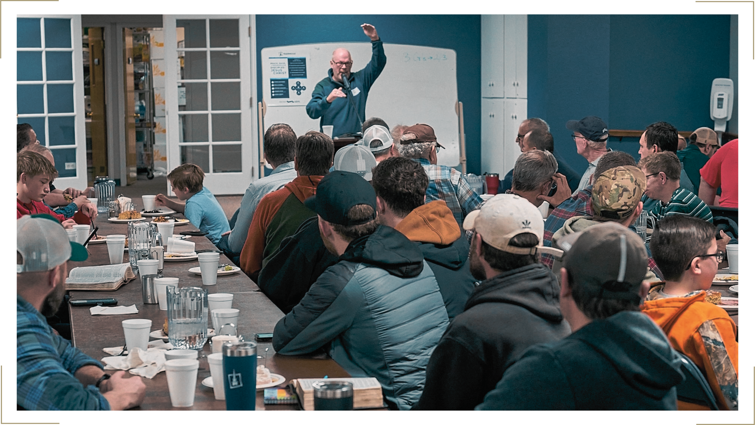 A man giving a presentation to a large group of people seated at a long table during a workshop or conference. The audience includes both children and adults, some wearing caps and casual clothing, eating and drinking while listening.