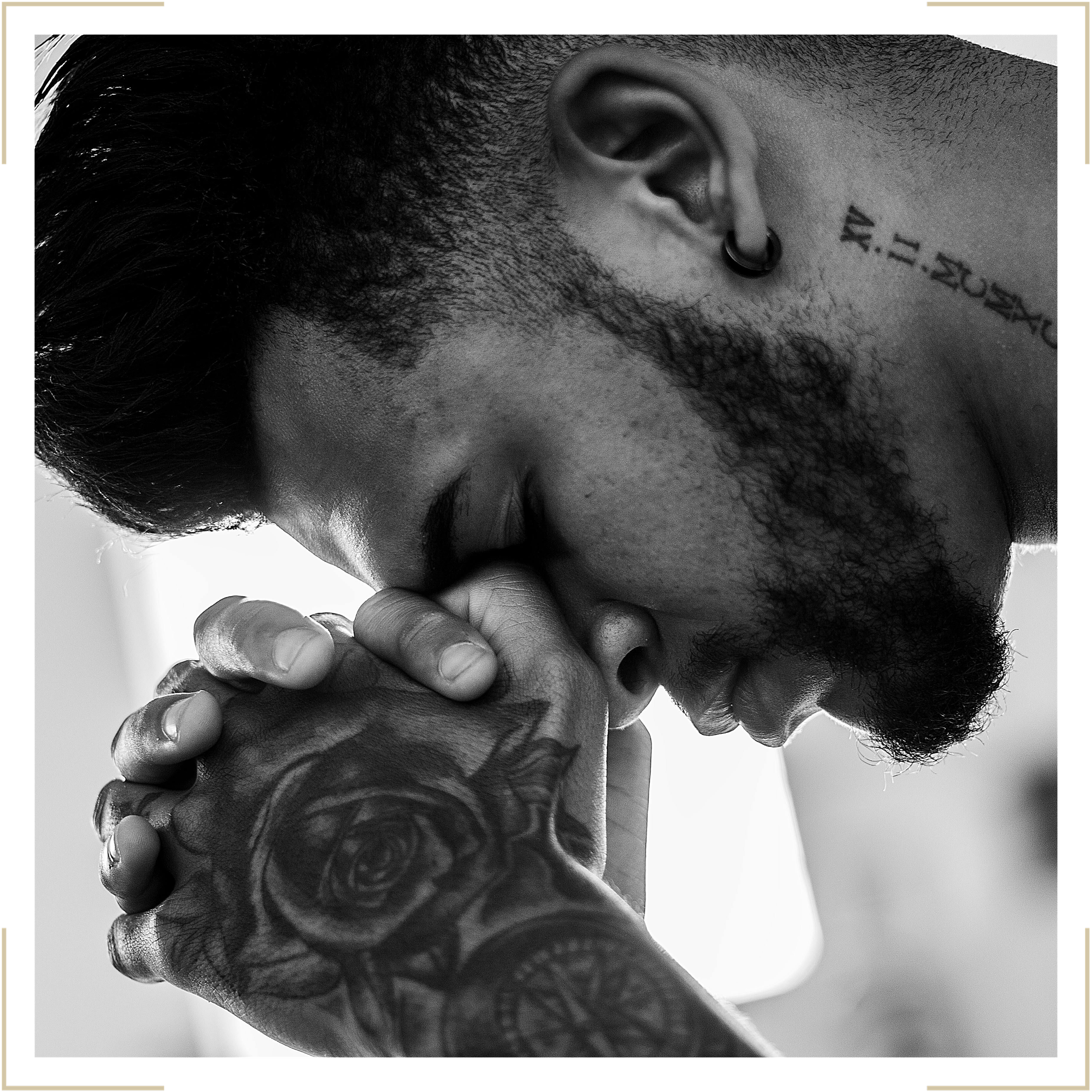 Black and white close-up photo of a man with closed eyes, a beard, tattoos on his neck and arm, and wearing an earring. His hands are clasped together near his face.