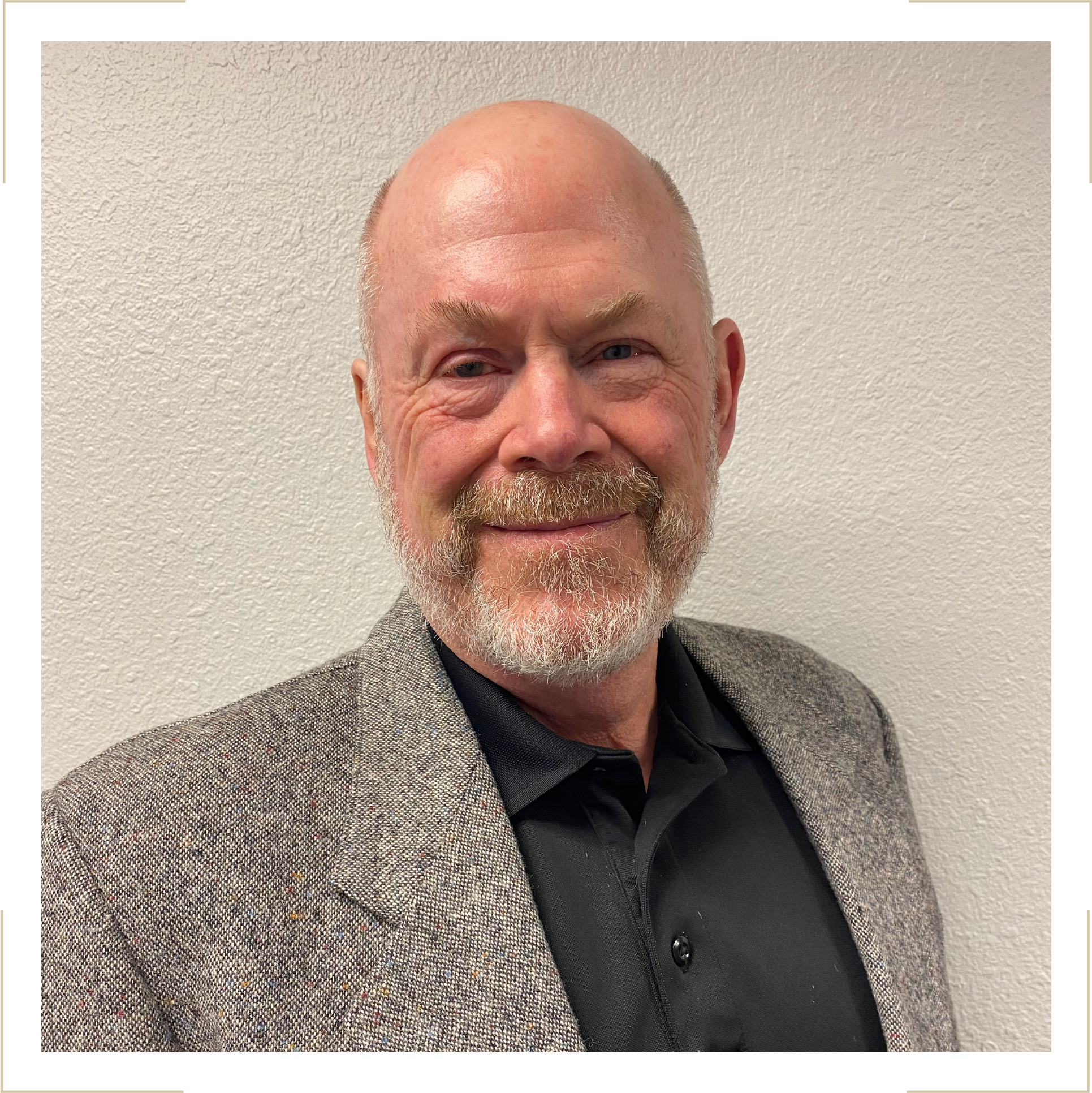 A middle-aged man with a beard and bald head, smiling, wearing a black shirt and a gray blazer, standing against a white textured wall.