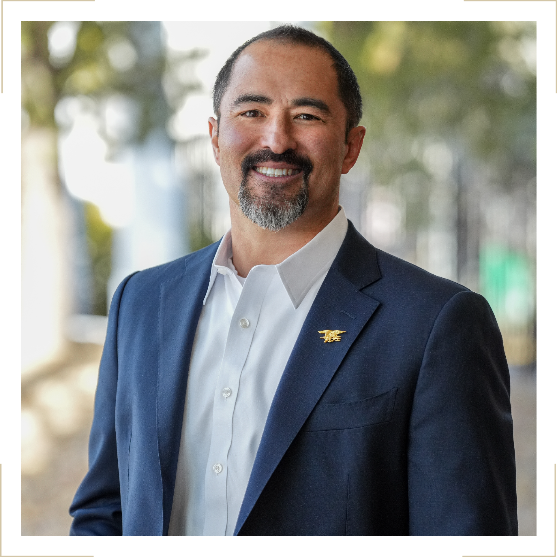A man with a beard and mustache, wearing a navy blazer with a gold lapel pin, a white dress shirt, standing outdoors with blurred trees in the background, smiling at the camera.
