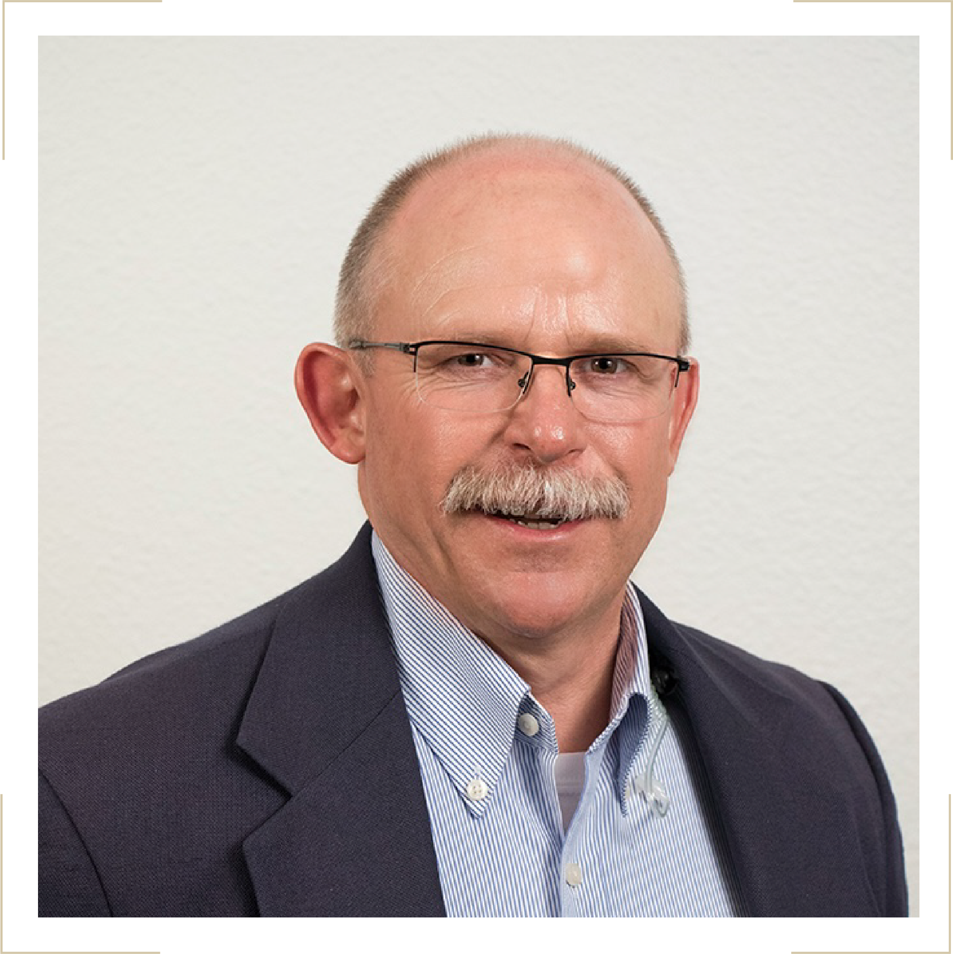A middle-aged man with a bald head, glasses, and a mustache, wearing a navy blazer over a light blue striped shirt, smiling in front of a plain white background.