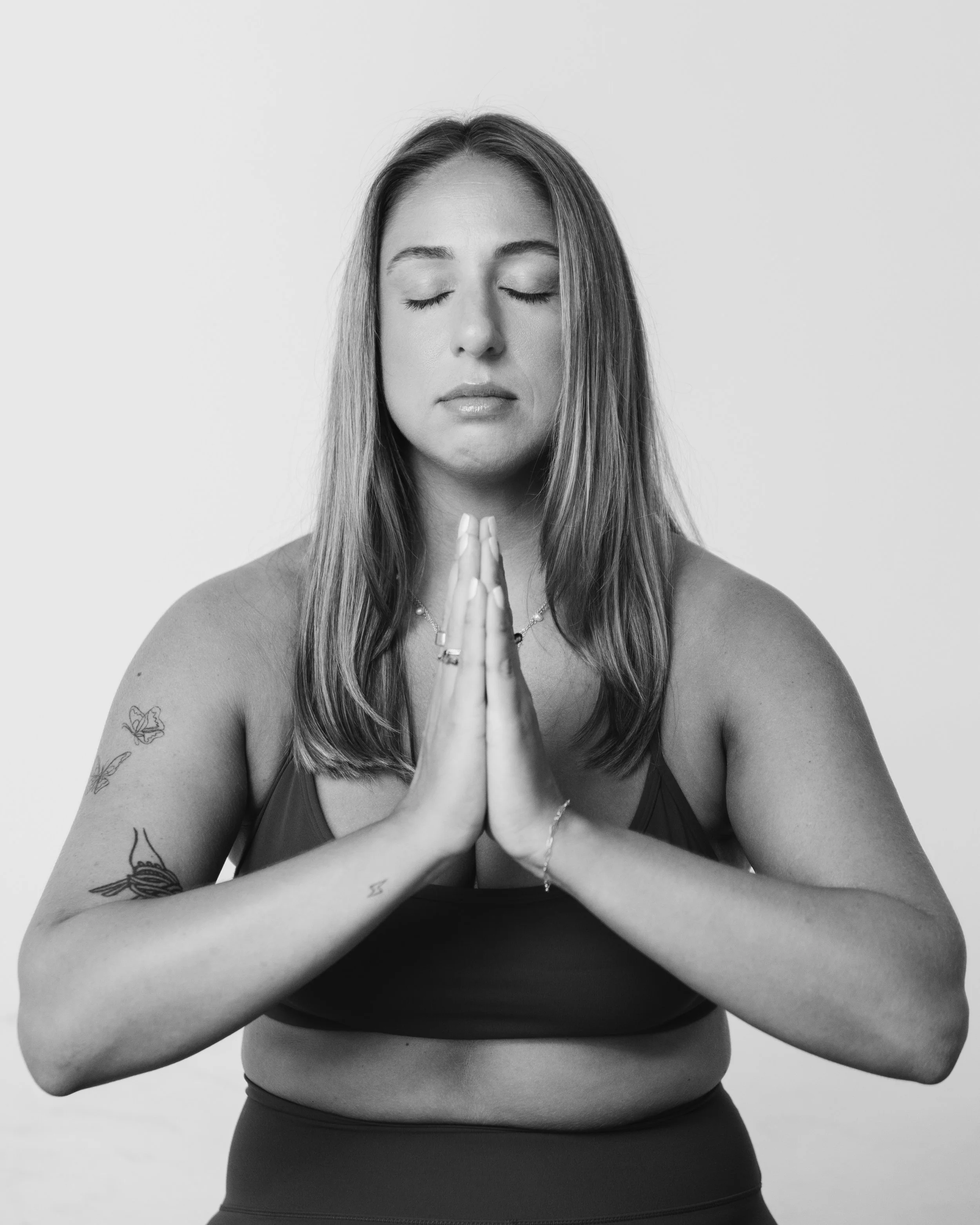 A woman with closed eyes in a meditation pose, palms together near her chest, tattoos on her arms, wearing a sports bra, against a plain background.
