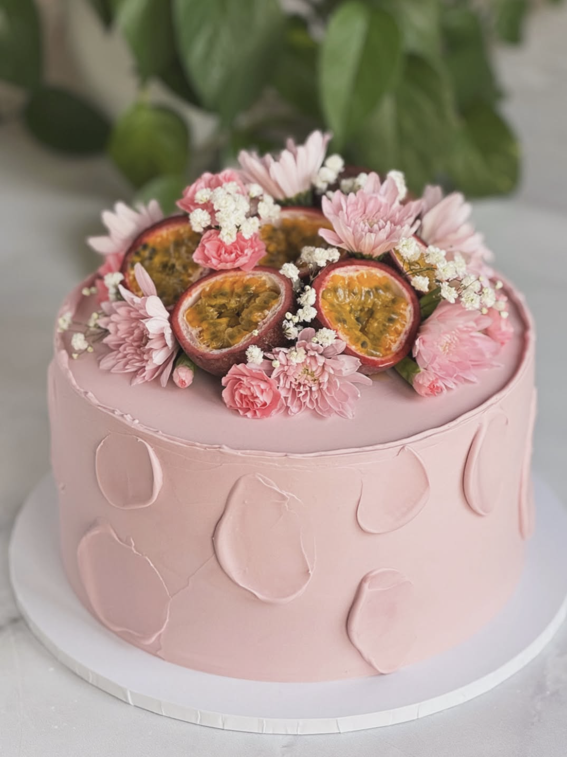 Pink cake decorated with passion fruit and pink flowers on top, with a backdrop of green leaves.