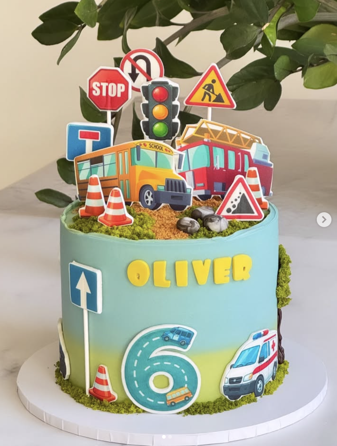 A colorful birthday cake decorated with toy traffic signs, a school bus, a fire truck, an ambulance, and traffic cones. The cake has the name 'Oliver' and the number '6' on it, indicating a 6th birthday celebration.