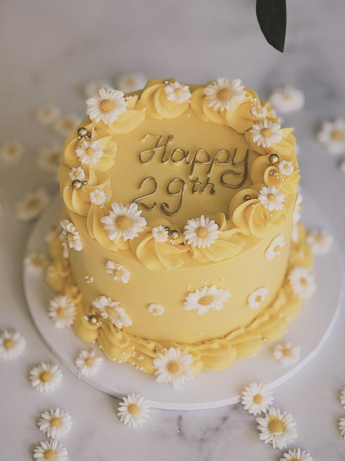 A two-tiered yellow birthday cake decorated with white daisies and gold accents, with the message 'Happy 29th' written on top.